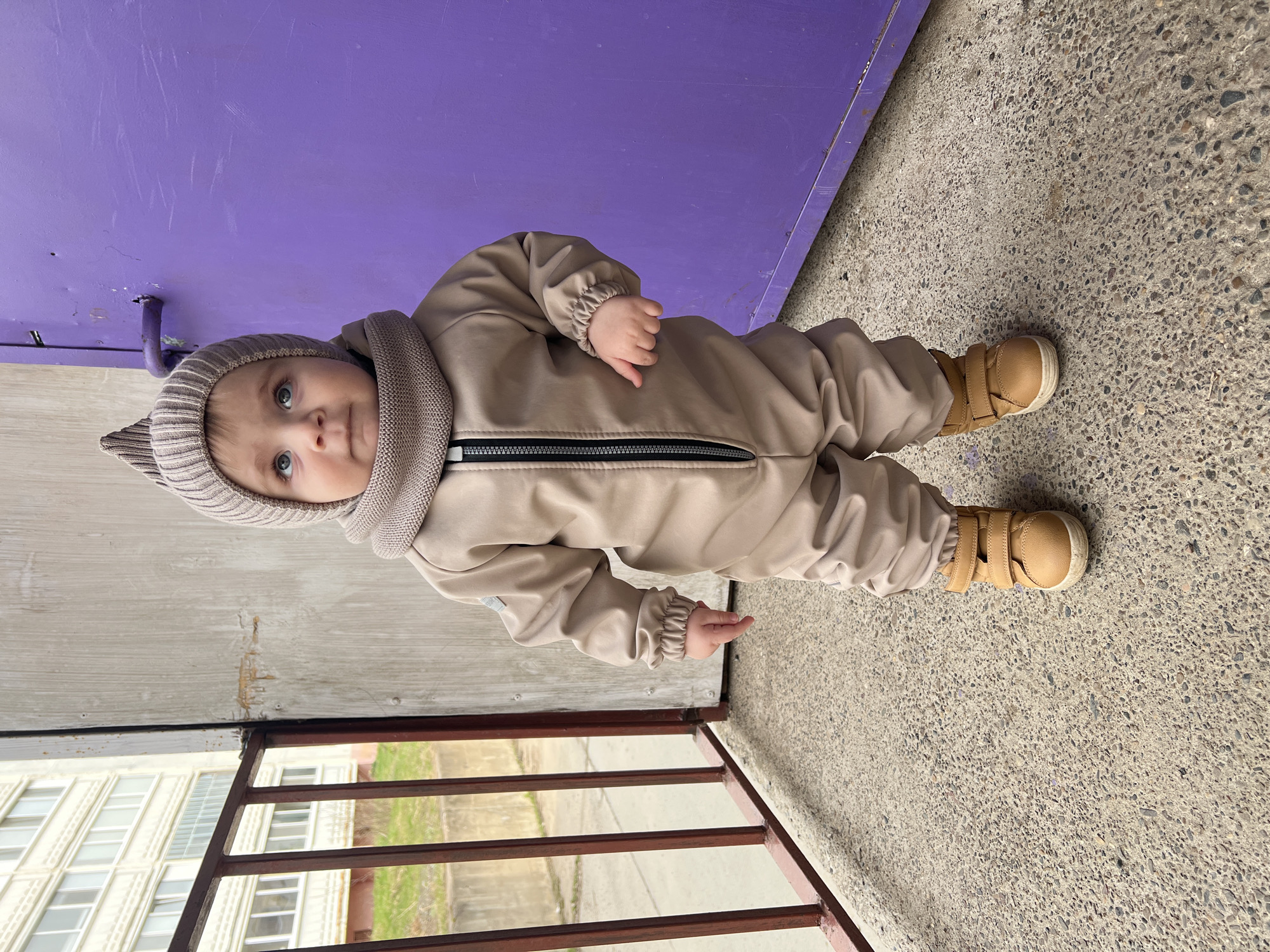 A baby is standing outdoors in Noviy, Russia. The baby is wearing a beige jumpsuit, a beige hat with ear flaps, and beige shoes with Velcro straps. The baby is standing on a textured concrete surface next to a purple wall and a metal railing. A building with white windows is visible behind the railing.