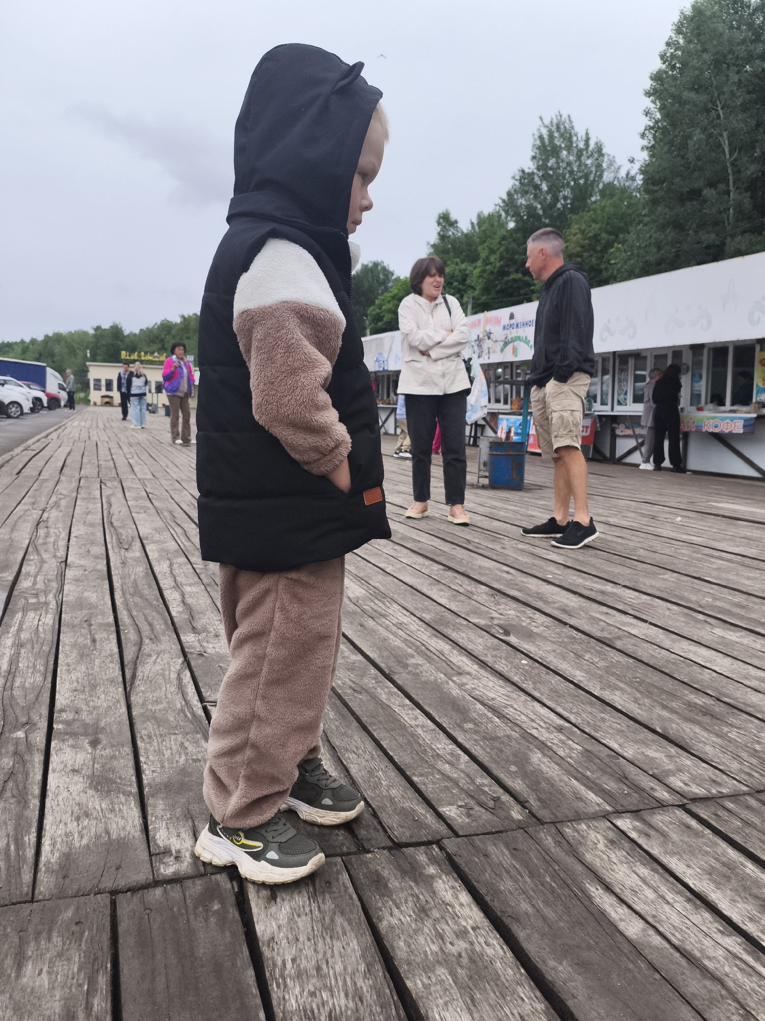 The image captures an outdoor scene on a weathered wooden boardwalk in Nizhnie Yaki, Russia, under an overcast sky. In the immediate foreground, a young blonde-haired child, positioned slightly left of center, faces right in profile, with hands in pockets. The child wears a black hooded jacket featuring small ear-like protrusions on the hood, a cream and light brown fleece mid-section, and matching light brown fleece pants. Dark grey athletic shoes with prominent white soles and yellow accents are visible on the child's feet.
In the midground, a woman with short dark hair, dressed in a light-colored jacket and dark trousers, stands with her arms crossed, appearing to engage in conversation with a man beside her. The man, with short dark hair, wears a dark hooded sweatshirt and light beige shorts, standing with his hands in his pockets. Further back, a series of low white commercial structures extends horizontally across the frame. These buildings feature multiple stalls and display signage in Cyrillic, with "МОРОЖЕННОЕ" (Ice Cream) clearly visible on one sign and "КОФЕ" (Coffee) on another to the right. Several other individuals are observed walking or standing near these structures and further down the boardwalk. Green deciduous trees are visible behind the buildings, forming a natural backdrop. To the far left in the distance, a parking area with several vehicles is present. The wooden boardwalk planks show signs of age and wear.
