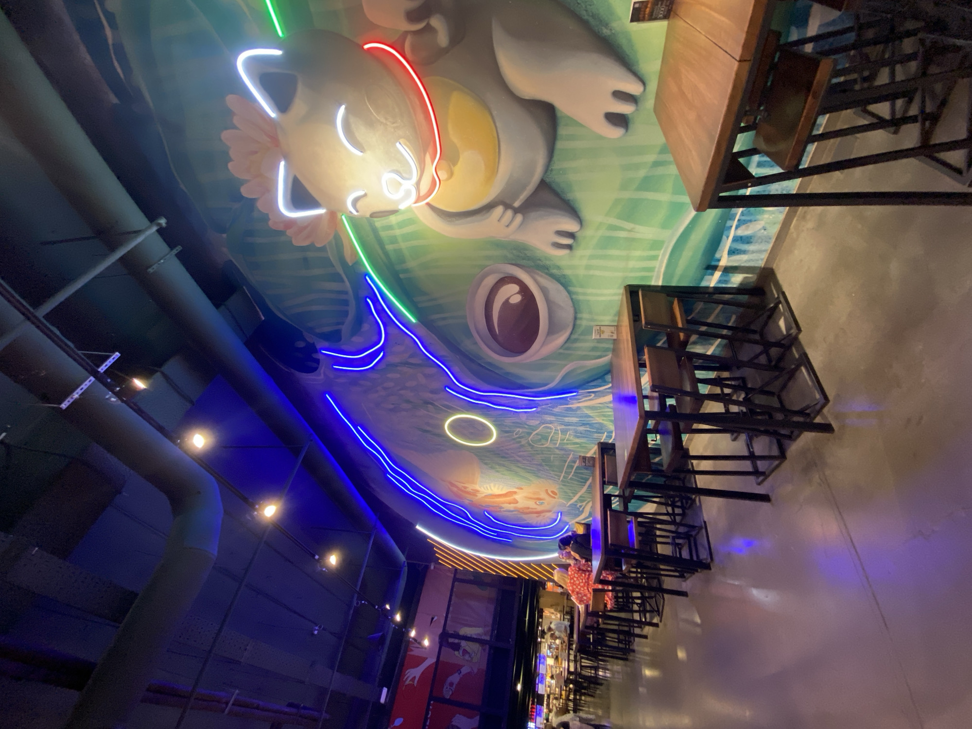 This image captures the interior of a restaurant with a playful, cartoon-themed mural on the ceiling. The mural features a large, stylized cartoon character, possibly a cat or a bear, depicted with a friendly expression and outlined in neon lights of red, white, and green. Below the character, a large circular shape with an eye-like pupil is also visible, part of the mural's design. Neon blue lights trace a wave-like pattern across the ceiling, complementing the underwater or aquatic theme suggested by the mural's background.
In the foreground, to the right, several wooden tables with metal frames and chairs are neatly arranged. These tables appear to be set up for dining. A few people are visible in the background, including one person in a red and white patterned dress standing near the tables. The lighting in the restaurant is dim and atmospheric, with the neon lights providing vibrant accents against the darker tones of the industrial-style ceiling pipes and the concrete floor.
The scene suggests a lively and modern dining establishment, possibly a cafe or a themed restaurant, designed to be visually engaging. The image is taken from a low angle, looking upwards towards the ceiling mural and the dining area. No specific time of day or weather conditions are discernible from the indoor setting. There is a visible menu board attached to one of the tables. The overall mood seems relaxed and inviting.