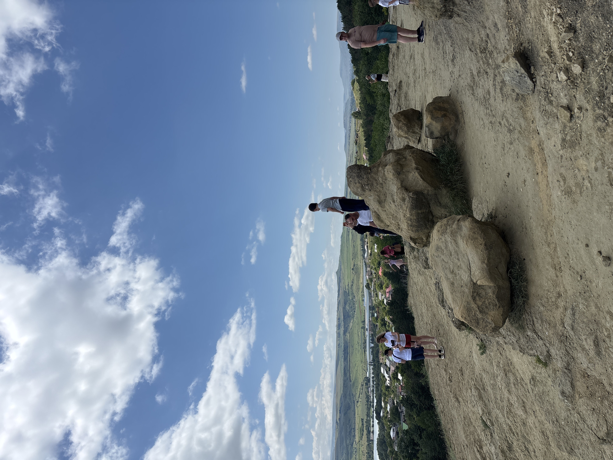 A group of people are gathered on a rocky, dusty hillside overlooking a valley with a town, a body of water, and rolling green hills under a bright blue sky with fluffy white clouds. The people appear to be tourists, dressed in casual summer clothing. Some are standing and looking at the view, while others are talking amongst themselves. One person in the foreground is a man wearing a beige t-shirt and turquoise shorts. To his left, a group of four people are standing near some large boulders; two adults and two children. Further into the scene, other individuals are visible, also appearing to be enjoying the scenic overlook. The location is described as a hillside in the city of Mirny, Russia.