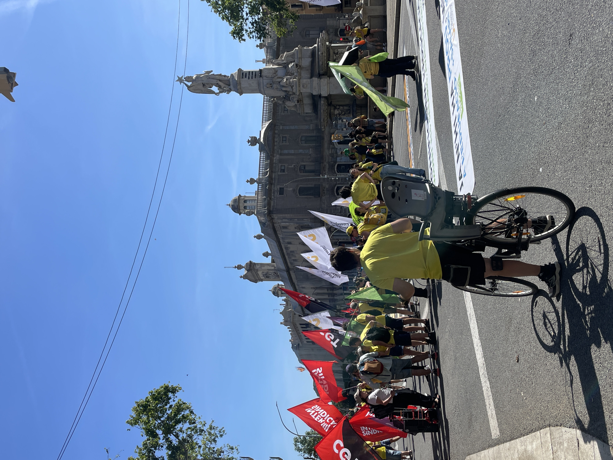 A vertical, eye-level photograph shows a public demonstration or parade in a city. A person rides a bicycle in the right foreground, obscuring a portion of the scene. They are wearing a yellow shirt and dark shorts. A child seat is mounted on the back of the bicycle.
Behind the cyclist, a crowd of people is marching in the street, holding red and white flags and wearing yellow shirts. The flags have lettering and symbols on them.
In the background, a large stone building with spires and statues is visible. A tall monument featuring a statue is situated in front of the building. The sky is blue and clear.
The location is a city street, likely in Italy, based on the flags and architectural style. The city and specific location are unknown.