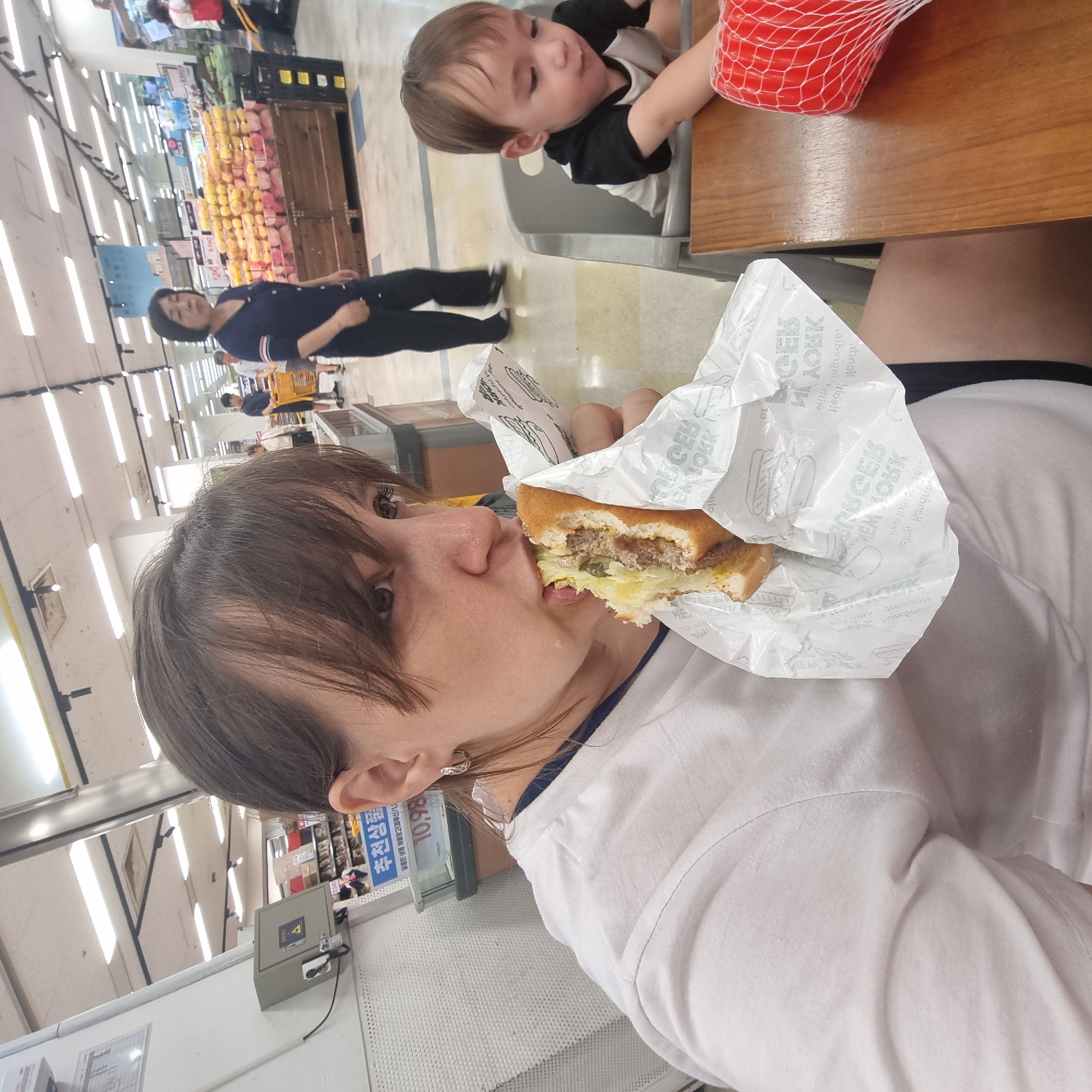 In a well-lit supermarket in Wonju-si, South Korea, a woman is captured in a close-up, mid-bite of a hamburger. She is wearing a white t-shirt and has dark hair with bangs. To her right, a young child sits in a high chair, facing away from the camera, looking down at something on the table. The child wears a black and white shirt. The background reveals aisles of products, including fruits displayed on wooden crates. Other shoppers can be seen in the distance, including a woman in a dark blue top and dark pants, and a person pushing a yellow shopping cart. A sign with Korean text is visible on a shelf. The overall scene suggests a casual outing to a grocery store.