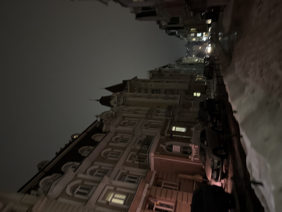 A dimly lit urban street scene at night. Elaborate buildings line the left side, featuring multiple stories, arched windows, and ornate architectural details. Several cars are parked along the street. Snow is piled up on the right side of the road. Distant streetlights illuminate the scene further down the street, adding depth. The sky is a dark, overcast gray.