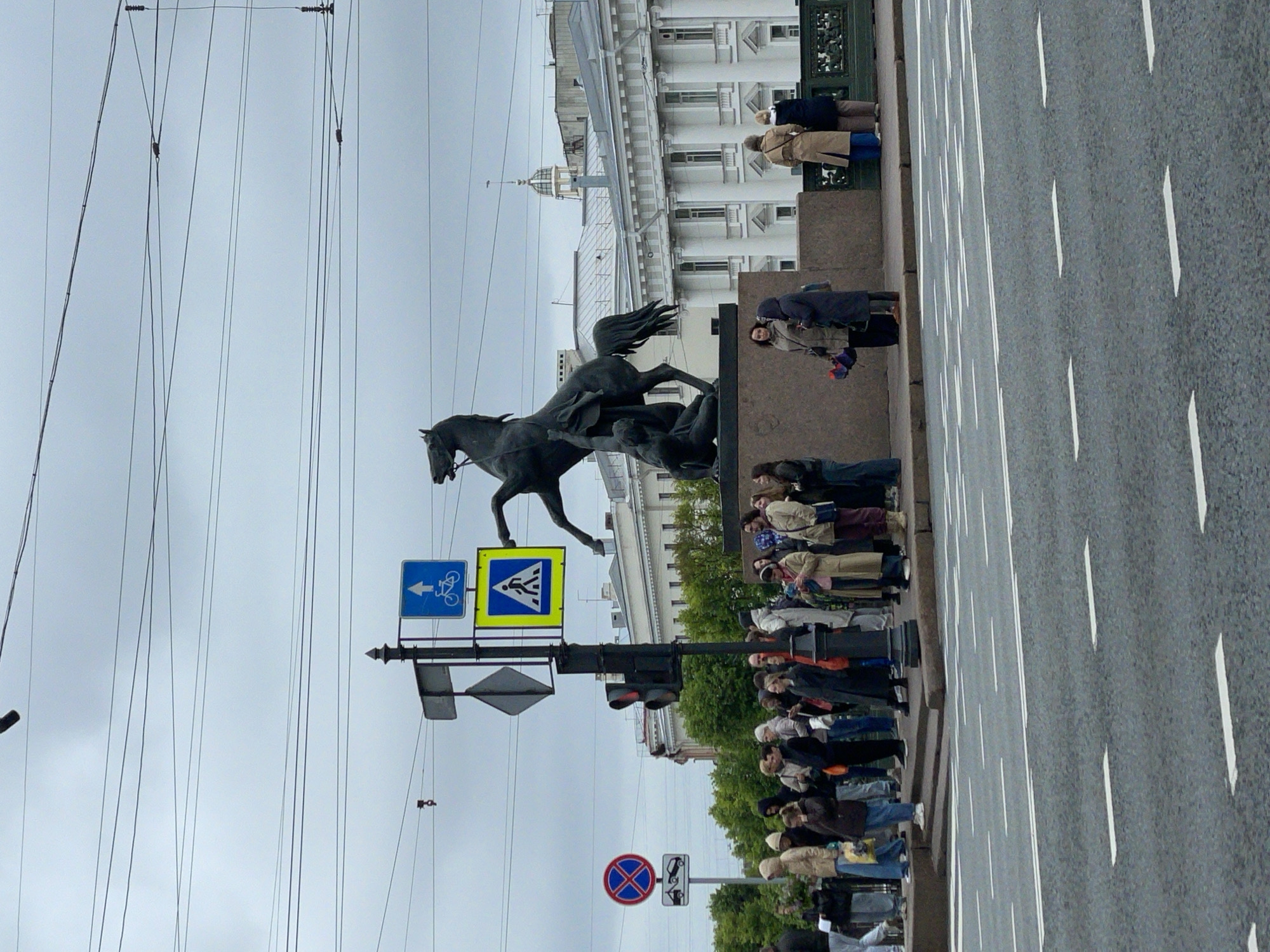The image depicts the Anichkov Bridge in Saint Petersburg, Russia, known for its horse sculptures. The bridge spans the Fontanka River, visible as a road with white lane markings on the right. On the left stands one of the iconic horse sculptures. Several people are present, seemingly tourists or locals, standing on the sidewalk along the bridge. Signage for pedestrians and cyclists is visible on a pole near the sculpture. Overhead, power lines crisscross the sky. The backdrop includes a building with white columns. The sky is overcast.