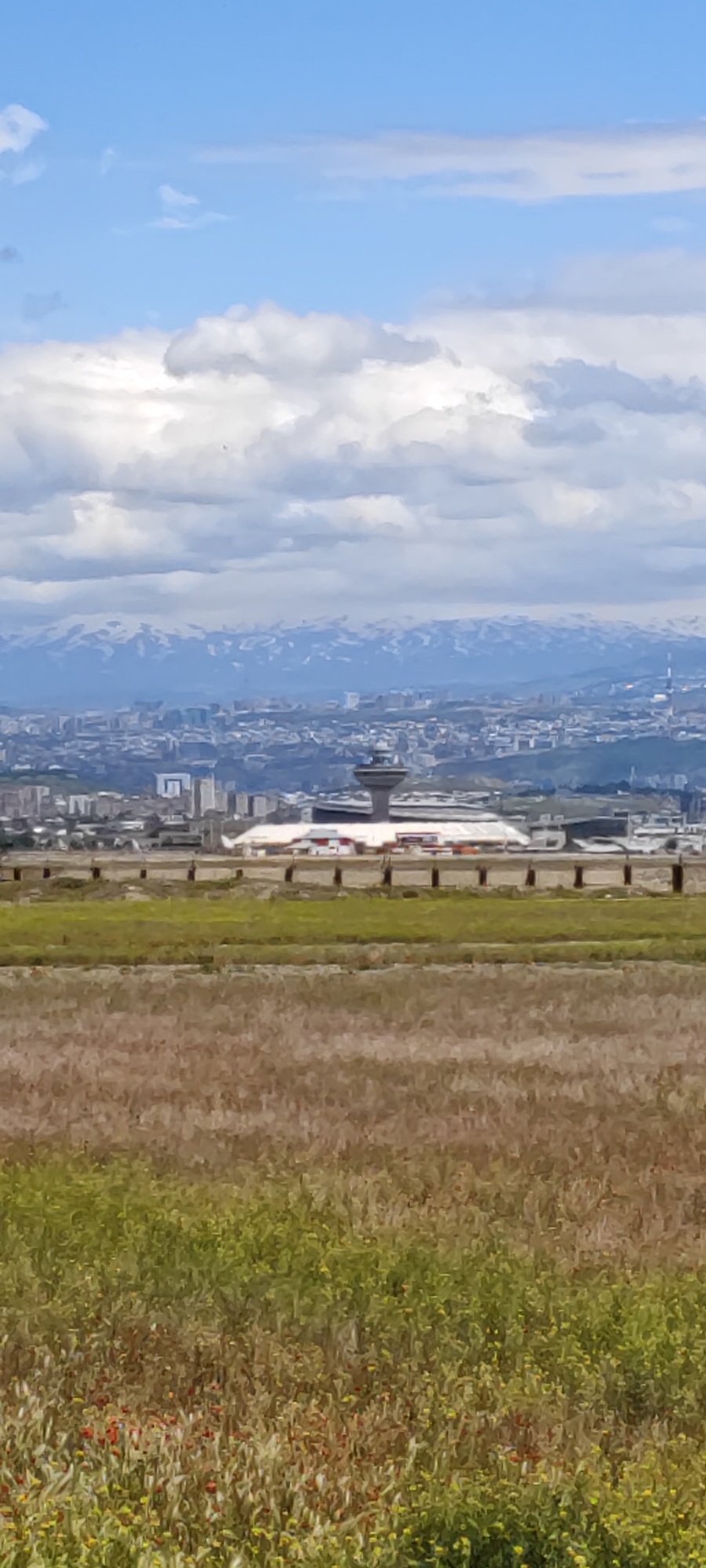 A long shot depicts the "Zvartnots International Airport" in Arevashat, Armenia. The airport features a control tower and terminal. In the background, a cityscape fades into snow-capped mountains under a sky with patchy clouds. In the foreground, there is a field with green and brown vegetation.