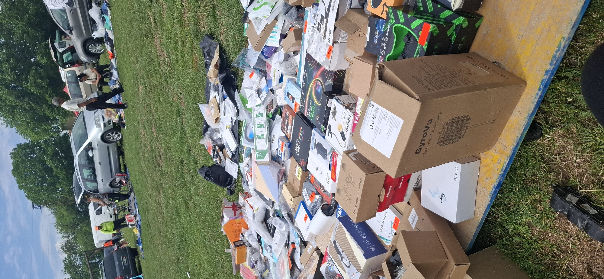 The image captures an outdoor market scene in a grassy field in Gorno Sahrane, Bulgaria, under a partly cloudy sky.
In the foreground, a weathered wooden platform, featuring yellow and blue paint, holds a substantial, disorganized pile of packaged consumer goods. These items are primarily in cardboard boxes and plastic packaging, suggesting new or open-box merchandise. Visible product names include "GyroVu USB Crystal Touch Lamp," "DISwoe," "Wireless Controller For PS4," "3D ILLUSION LAMP," "LED BOXKU," "MP3 Music Player," "USB Cabinet Light," and "Horizontal Employee Badges." Several items display price tags in Bulgarian Lev (лв), such as 20лв, 22лв, 25лв, and 95лв. Black plastic bags and other packaging are intermingled with the products.
The mid-ground features several parked vehicles on the grass, indicating vendor stalls. From left to right, these include a dark grey hatchback with license plate 3671W82, a white commercial van, a silver minivan (likely a VW Sharan) with its rear hatch open, a beige/tan minivan, and a dark grey SUV with its rear hatch also open.
Several individuals are present, engaged in market activities. Near the white van, a person wearing an orange cap and a bright green long-sleeved shirt stands beside a bicycle rack. Another person bends over near the silver minivan's open trunk. A man with grey hair, wearing a light-colored short-sleeved shirt and dark trousers, walks towards the right. Further right, a person is seated on the grass near a spread of clothes and other items.
The background consists of numerous green deciduous trees under a blue sky with scattered white clouds. A dark utility pole or similar structure is visible in the distance among the trees. The overall environment is an open, grassy area characteristic of an informal outdoor market or flea market.