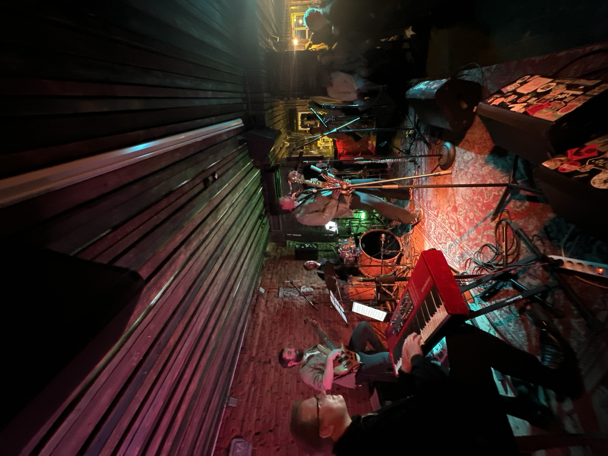 In Minsk, Belarus, a band performs on a dimly lit stage in what appears to be a bar or small venue. The band consists of at least five musicians. In the foreground, a keyboardist wearing glasses and a black top plays a red keyboard. To their right, a bassist with a beard, wearing a grey shirt and blue jeans, plays a bass guitar. Behind them, a drummer is seated at a drum set. Further back and to the right, a saxophonist in a light-colored jacket plays a saxophone. Another musician, possibly a vocalist or another instrumentalist, stands near the saxophonist. The stage is adorned with a patterned rug, and various musical equipment, including microphones, amplifiers, and speakers, are visible. The background features a brick wall and a wooden slatted ceiling, with colored stage lighting casting red and green hues across the scene. In the far right of the image, a crowd of people watches the performance.