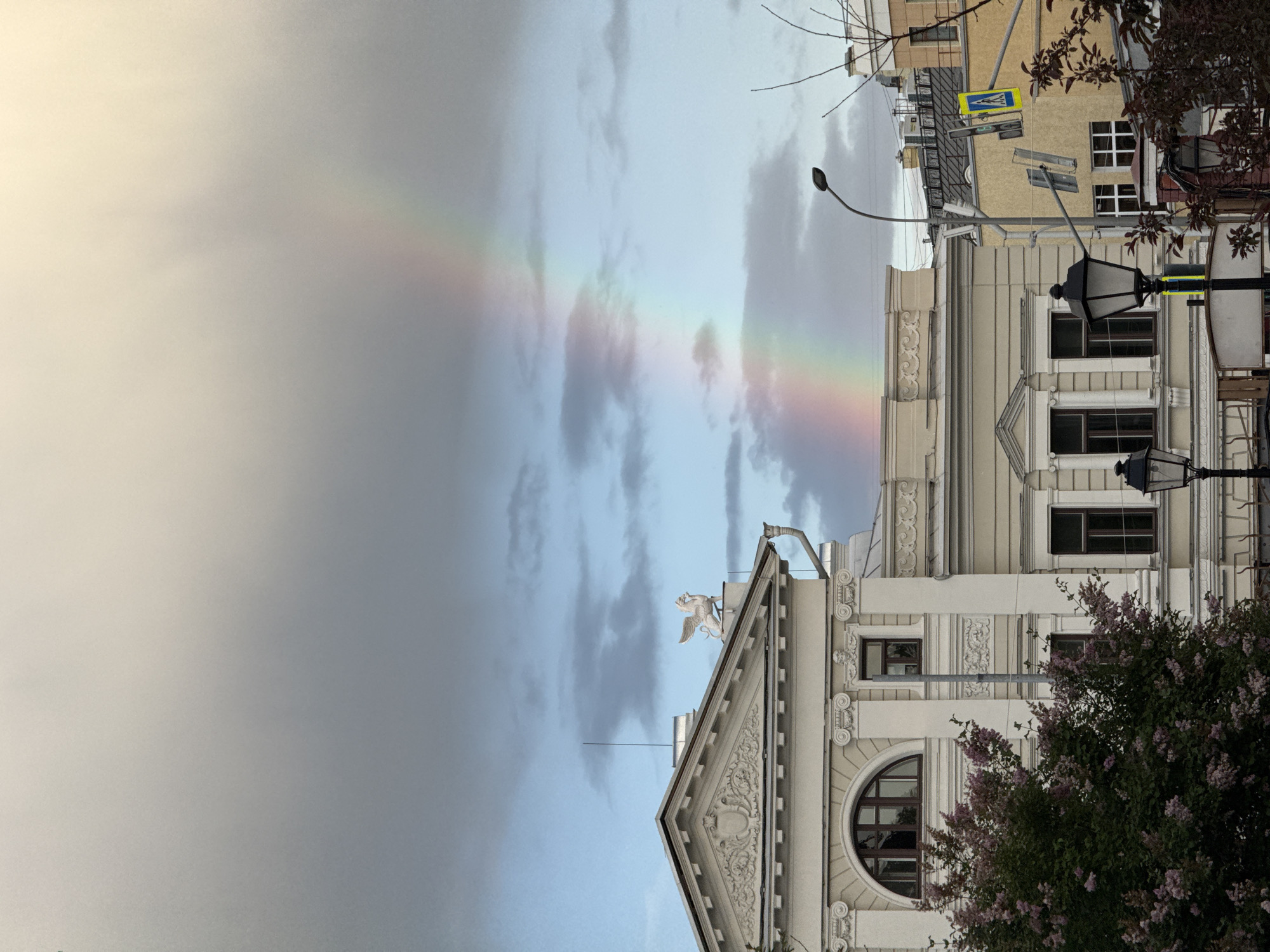 A vertical image shows a partial rainbow in the sky above Moscow, Russia. The rainbow arcs from the left side of the frame towards the right, disappearing behind a light-colored building. The architecture of the building is ornate, with decorative moldings and a statue of a winged griffin atop its triangular roof. Street lamps are visible on the right side of the image, with blooming lilac bushes partially obscuring the building's facade. A yellow building with a pedestrian crossing sign is visible behind the light-colored building. The sky is cloudy with patches of blue visible between the clouds.