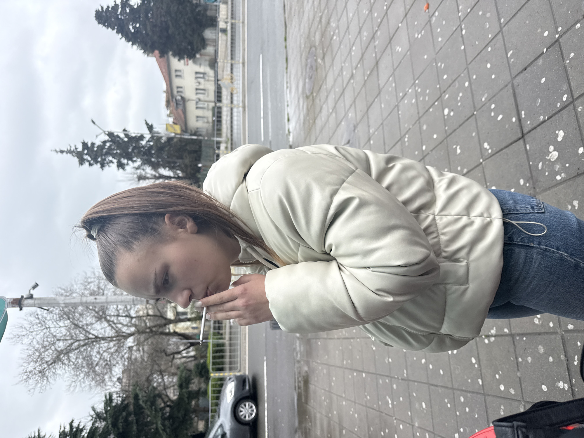 A young woman with brown hair pulled back into a ponytail is smoking a cigarette on a sidewalk. She is wearing a light-colored, hooded puffer jacket and blue jeans. The sidewalk is tiled with gray blocks dotted with white spots. Behind her is a street with a gray car parked along the curb, a few bare trees, and buildings in the distance. The sky is overcast. The buildings in the background are located in Burgas, Bulgaria.
