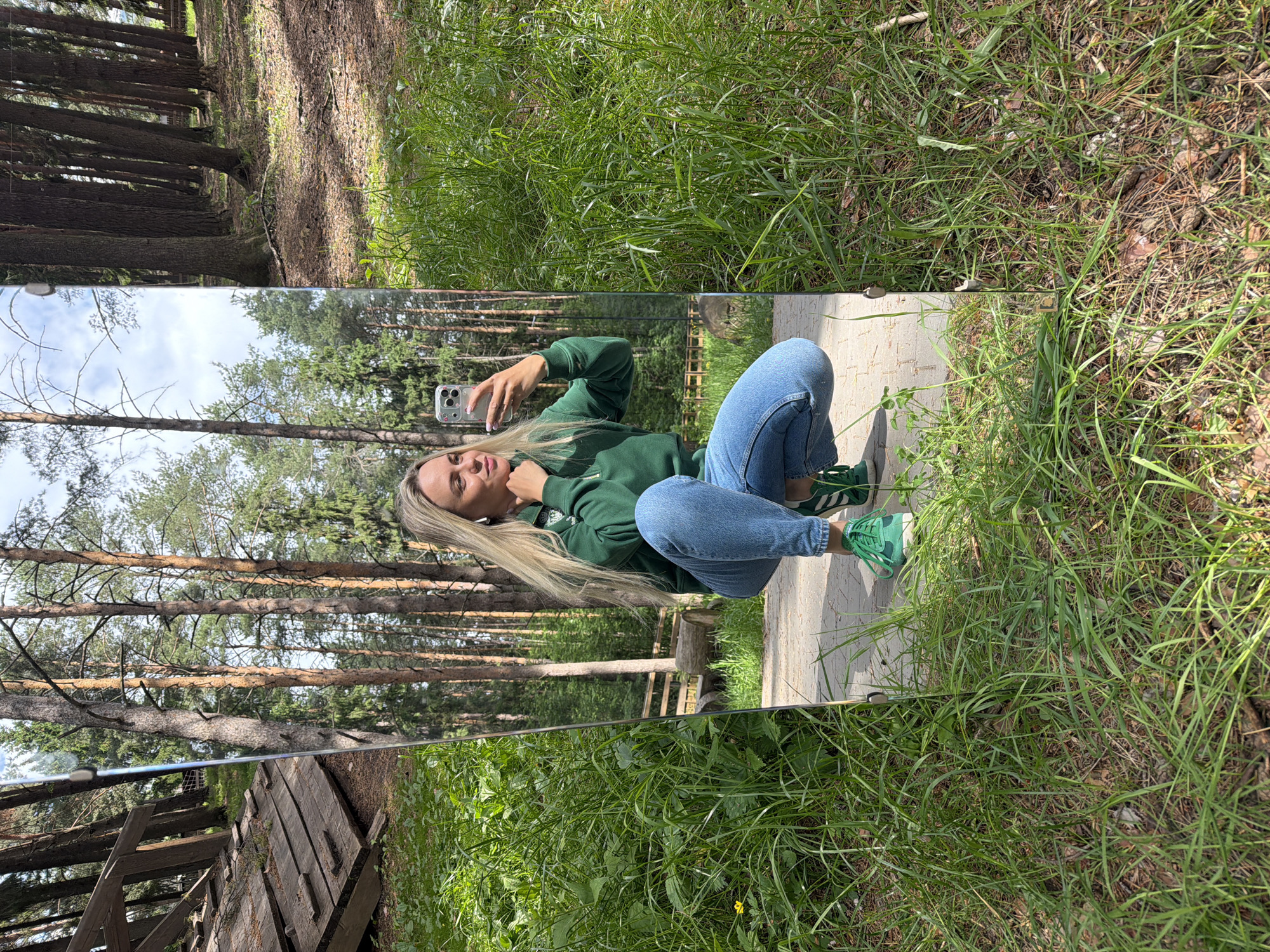 A blonde woman in a green hooded sweatshirt and blue jeans is squatting on a walkway in a grassy area near a forest. She is holding a smartphone and appears to be taking a selfie in a mirror that is positioned vertically in front of her. The mirror reflects the forest behind her, including trees and a wooden structure. The location is likely in a park or natural area, possibly near the city of Votkinsk, Russia.