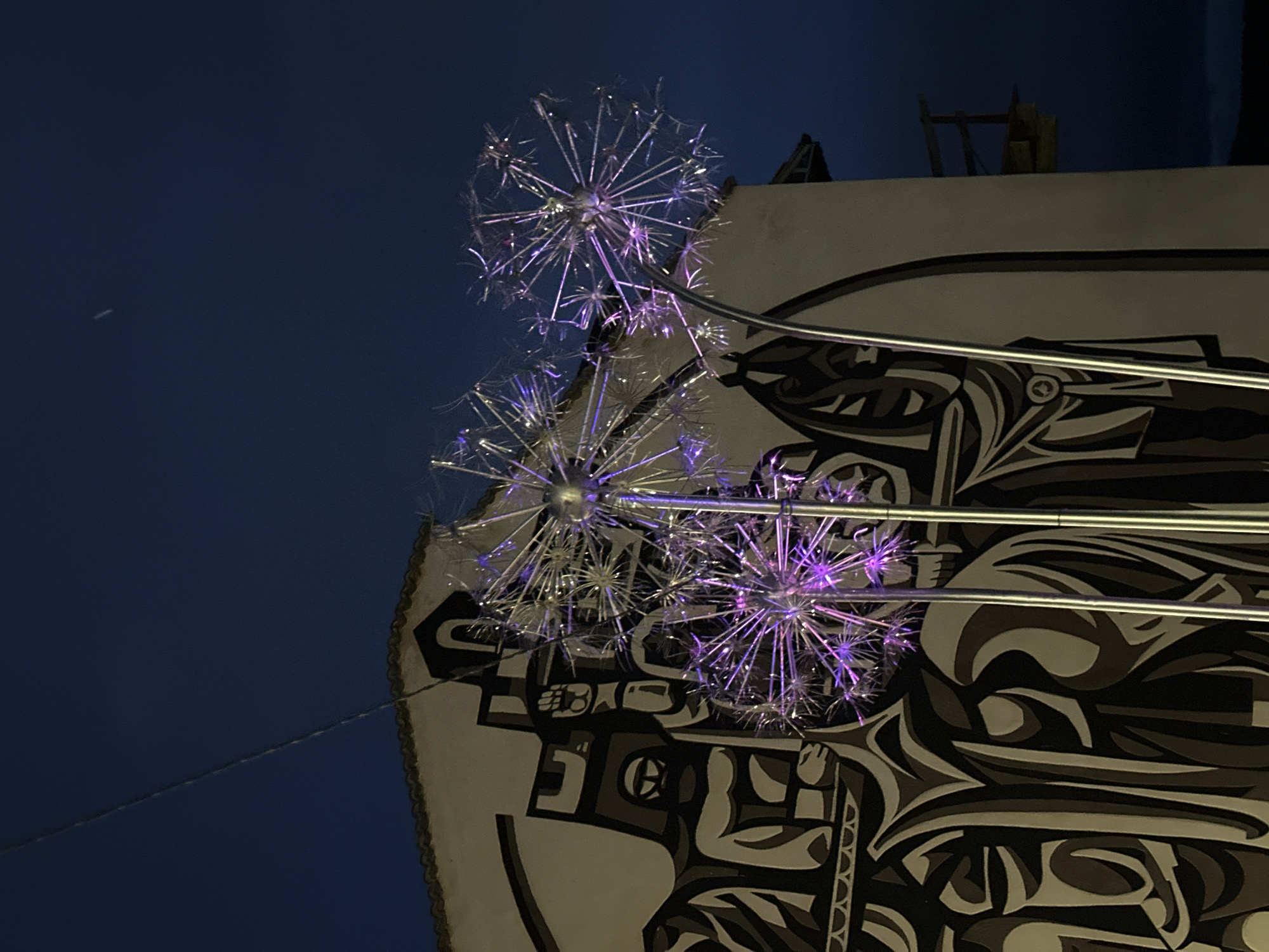 The image shows three illuminated dandelion-shaped light fixtures mounted on a building. The building has a white facade adorned with a gray and black mural depicting stylized figures and abstract shapes. In the background, the dark blue sky dominates the upper left portion of the image. A thin cable or wire stretches diagonally across the sky. Above the building, a glimpse of rooftop structures is visible. The scene is likely in an urban setting, given the architecture and the light fixtures. The exact location within Veliko Tarnovo, Bulgaria cannot be determined from this image.