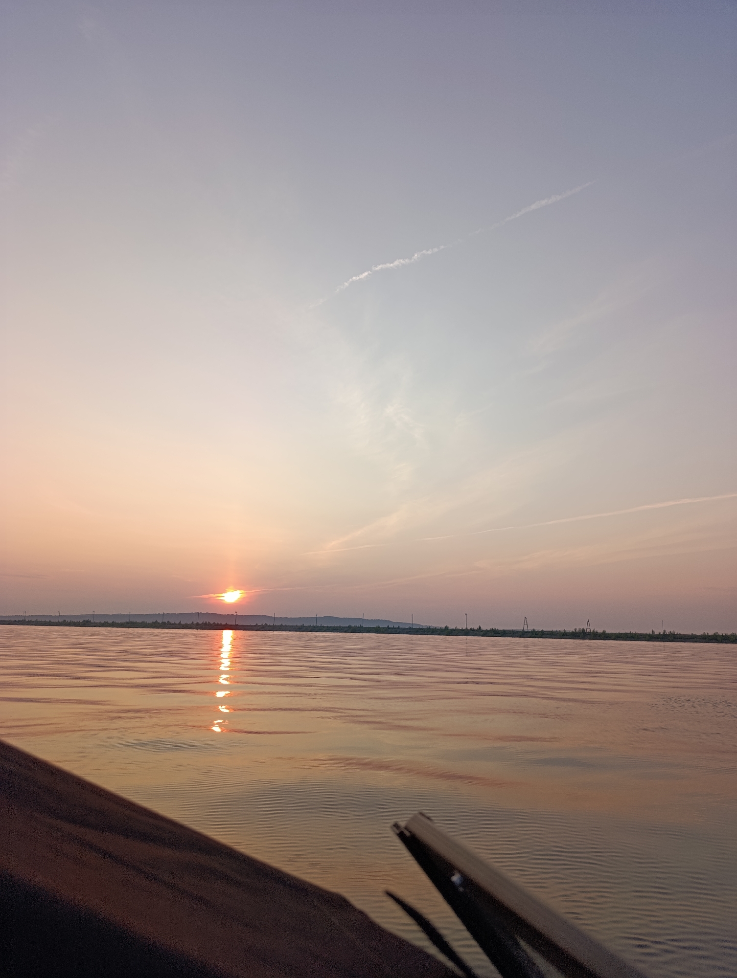 A low-angle, vertical, full shot captures a serene sunset over a body of water, likely the Kama River in Chaikovsky, Russia. The sun, a bright orb tinged with orange and yellow, is setting on the horizon, casting a shimmering reflection on the rippling water. The sky transitions from a pale lavender near the top to a soft, peachy orange near the horizon, with a thin contrail streaking across the upper portion. The distant shoreline is lined with a thin strip of trees and features a few scattered utility poles or pylons, indicating a populated area. In the foreground, the dark, wooden edge of a boat or vessel occupies the bottom left quadrant, with a blurred oar or paddle visible on the right, suggesting movement through the water. The scene is tranquil and devoid of human activity.