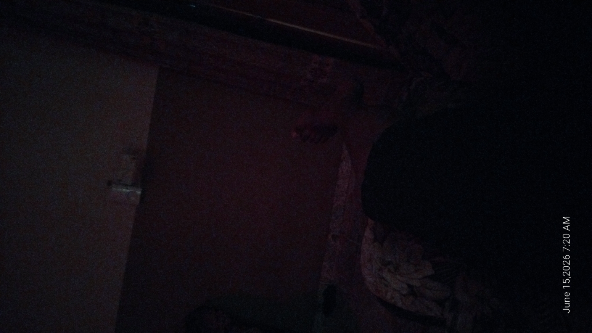 The image depicts a dimly lit interior space, likely a bedroom. The timestamp in the bottom left corner indicates June 15, 2026, 7:20 AM. A person's foot is visible on the right side of the frame, with toes pointing upwards. The rest of the person appears to be covered by bedding or blankets, suggesting they are lying down. The environment is dark, with limited visibility, but suggests a residential setting. No specific location within Howayi, Nigeria can be determined from the visual information provided. The scene appears to be a private moment, possibly captured upon waking.