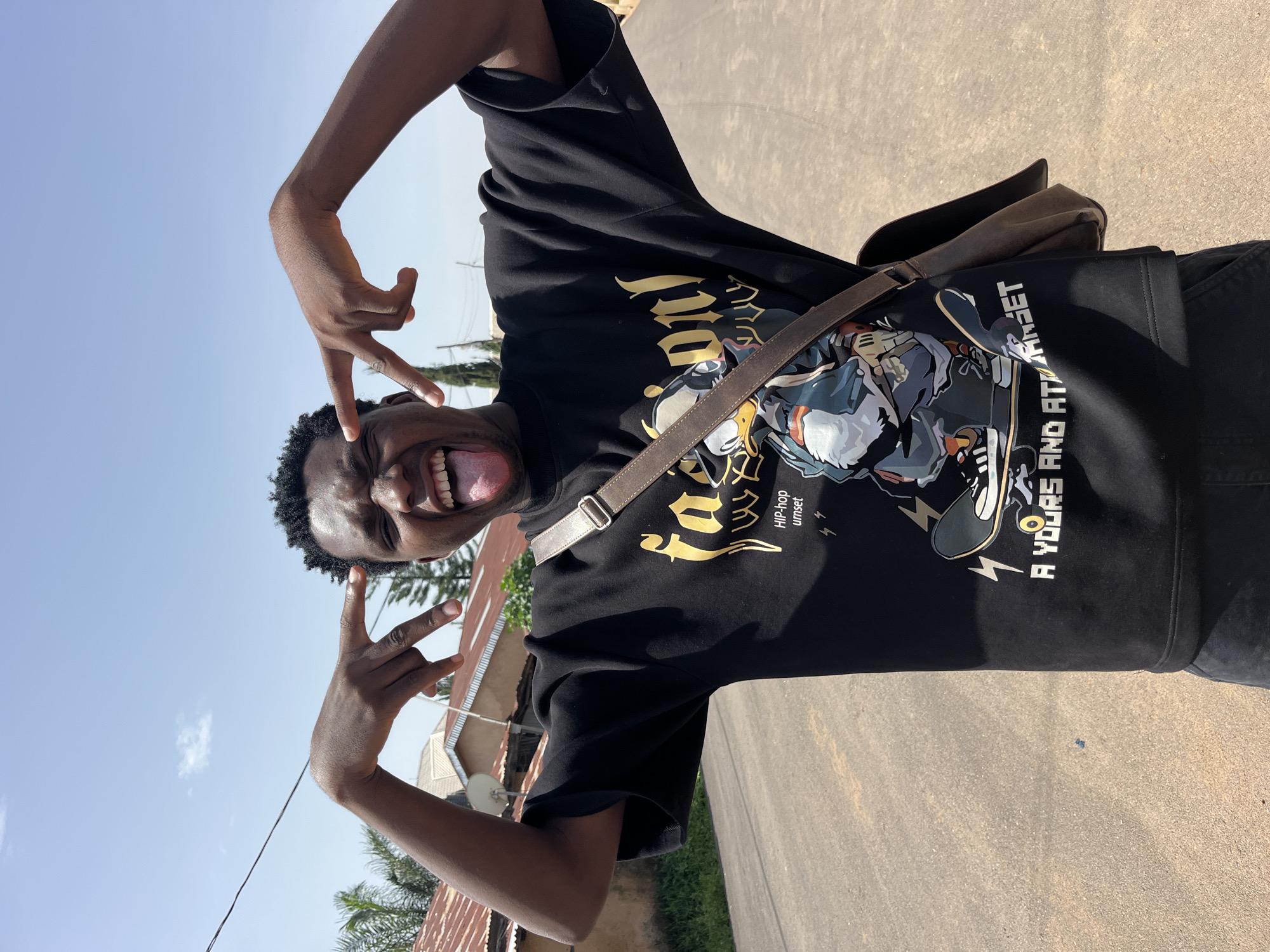 A young Black male stands in the middle of a paved road in a residential area. He is wearing a black t-shirt with a graphic print of a cartoon character on a skateboard and the text "FASION HIP-HOP UMSET A YOURS AND AT ARSET". He is making a peace sign with his left hand and a finger gun gesture with his right hand, while sticking his tongue out at the camera. He is wearing a brown messenger bag across his chest. In the background, there are houses with corrugated metal roofs and trees. The sky is clear and blue.