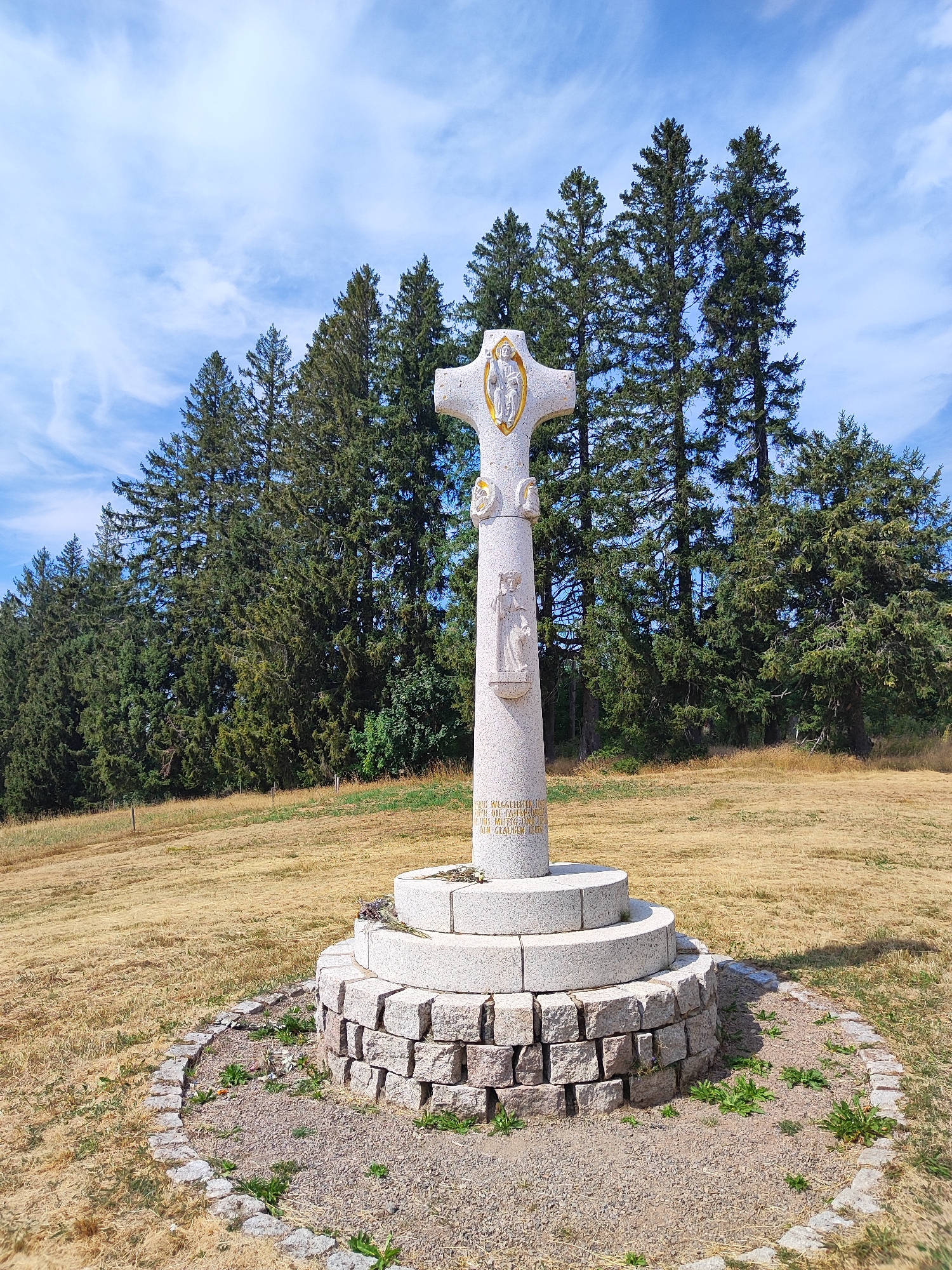 A stone monument in the shape of a cross stands in a dry, grassy field with a dense line of tall evergreen trees in the background. The monument is made of granite and has decorative carvings and inscriptions on it. It is surrounded by a circular base constructed of smaller stones. The sky above is blue with scattered white clouds. The scene appears to be located outdoors, likely in a rural or natural setting.