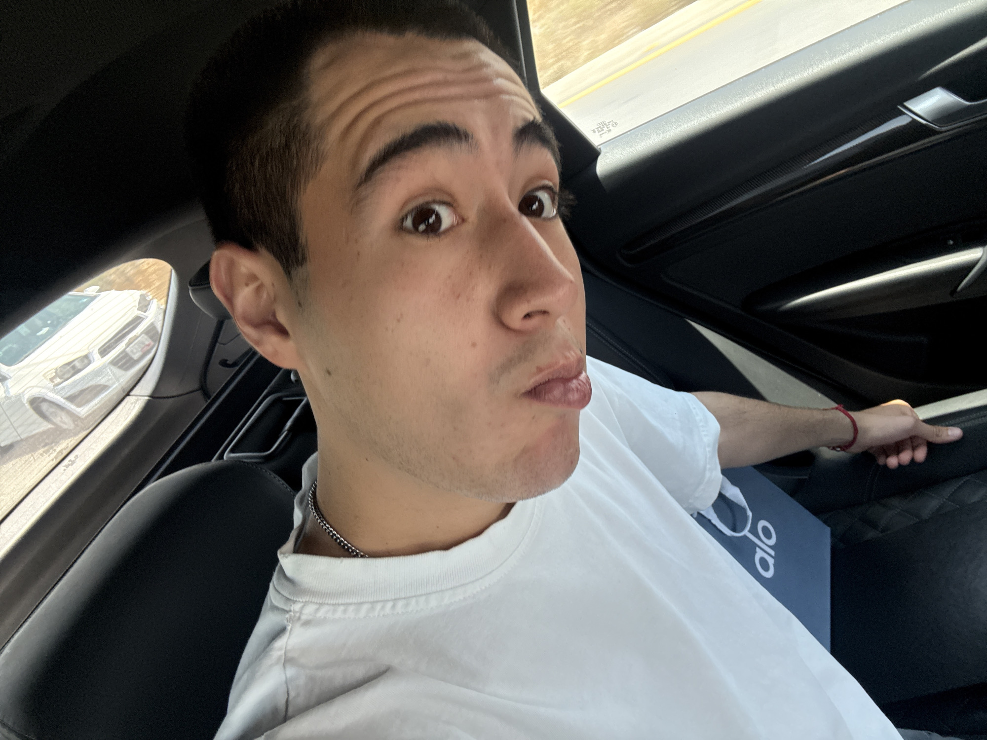 A young man takes a selfie from the passenger seat of a car. He has dark hair and eyes, and is wearing a white t-shirt and a silver chain necklace. His expression features pouting lips and wide, slightly surprised eyes, with visible wrinkles on his forehead. A red string bracelet is on his right wrist, which rests near the car's dark interior.
Next to him, a dark shopping bag with the white text "alo" is visible. The car's interior appears dark, possibly leather, with a quilted pattern on the seat beside the man. Through the car window, a road with a yellow lane line is visible, along with the reflection of another light-colored vehicle. The lighting suggests it is daytime. The scene is set in what appears to be a moving or parked car in the Barrio de Nuevo León, Mexico.