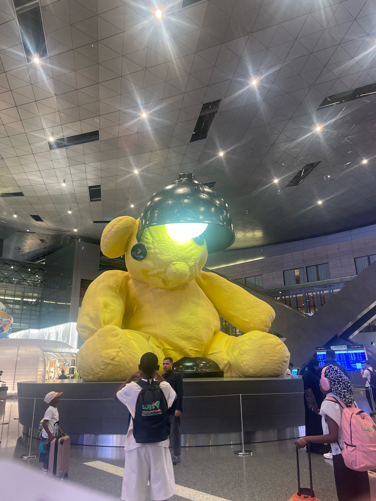 This image captures a large, vibrant yellow teddy bear sculpture with a dark green lampshade perched on its head, illuminated by a bright light. The sculpture is centrally located in a spacious, modern interior with a high, geometrically patterned ceiling featuring numerous light fixtures. Several people are present, including a young boy in a white t-shirt and a cap, pulling a pink suitcase, and a person in a white outfit with a backpack. A man in a suit stands behind them, and a woman with a pink backpack and patterned headscarf is visible on the right. The background shows a Louis Vuitton store and what appears to be an airport departure board. This scene is at Hamad International Airport in Doha, Qatar, known for its impressive public art installations.