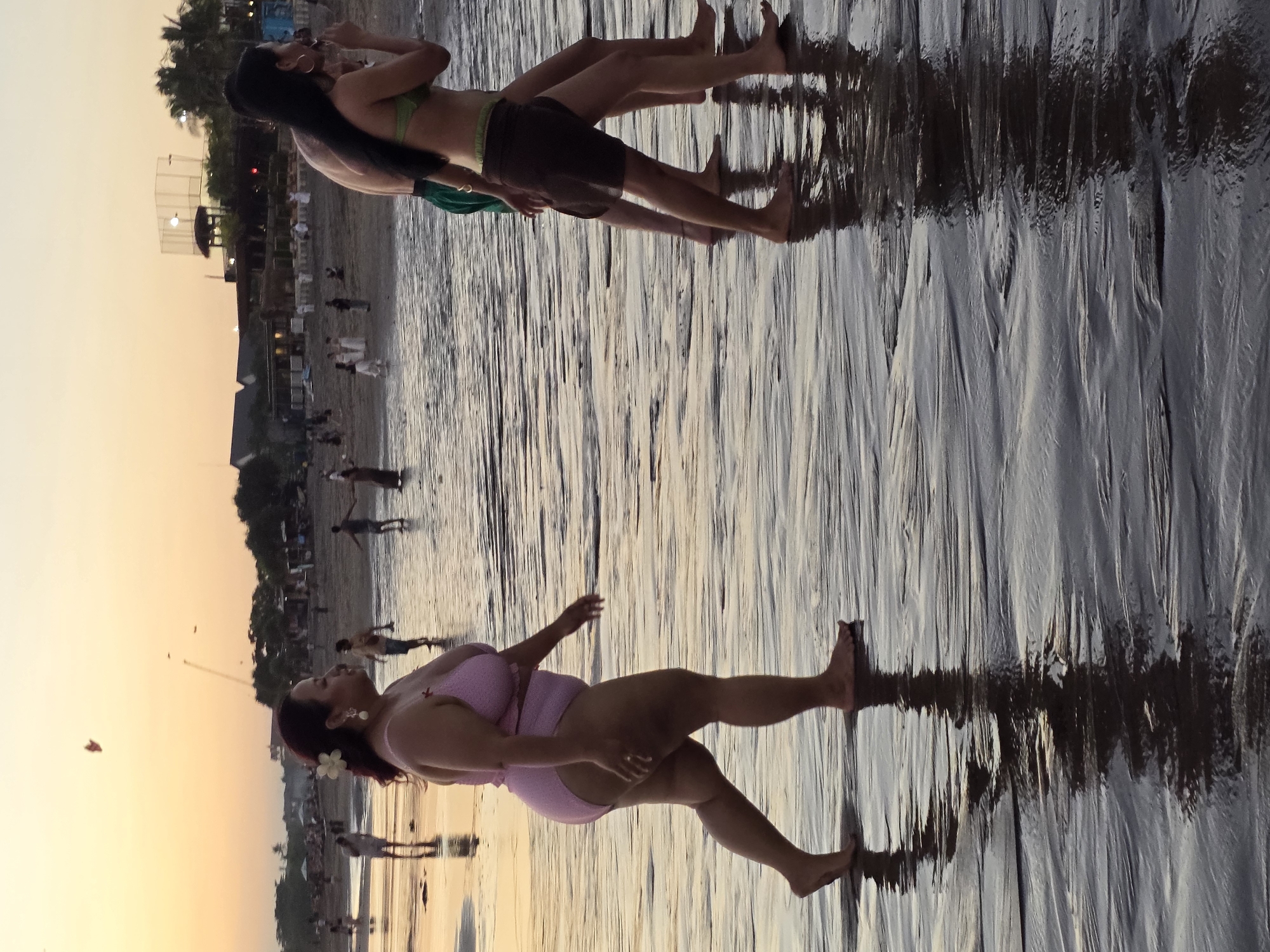 The image depicts a beach scene at sunset in North Kuta, Indonesia. In the foreground, a woman in a pink bikini stands on wet sand, looking to her left. She has a flower tucked behind her ear and is wearing earrings. Behind her, to the left and slightly out of focus, are two figures, one of whom is wearing a green bikini top and dark shorts. They are lying down and appear to be interacting. In the background, other people are visible on the beach, and there are some structures, possibly beach huts or tents. The sky is a soft gradient of orange and yellow, indicating the time of day. The wet sand reflects the sky and the figures, creating an elongated, mirrored effect. The overall atmosphere suggests a relaxed evening on the coast.