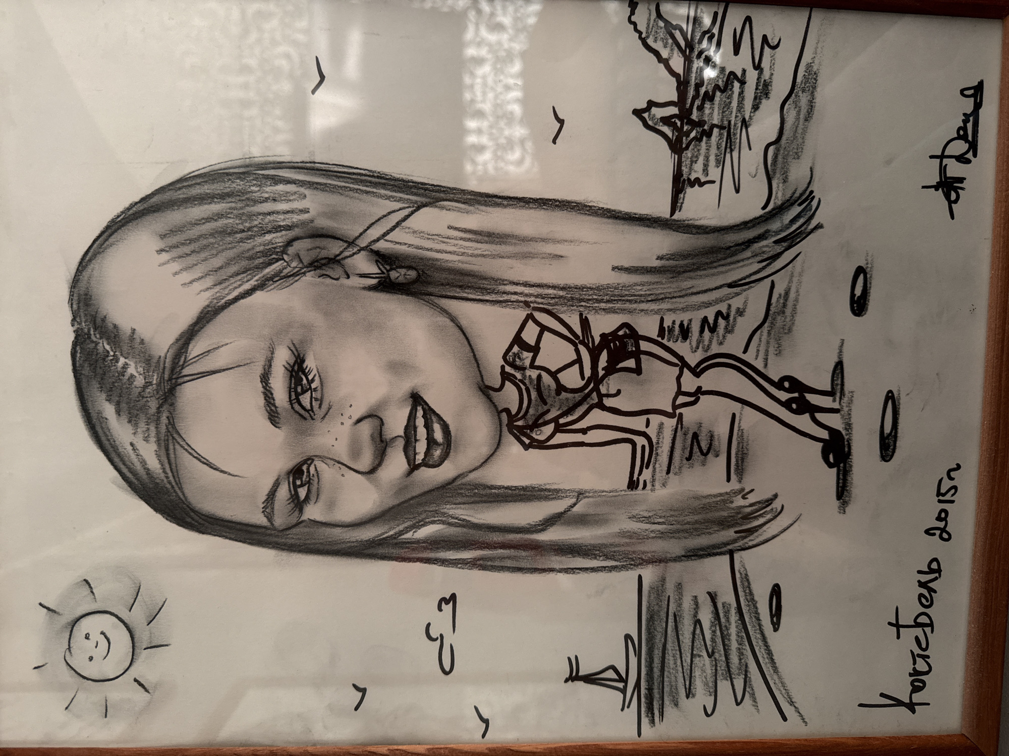 The image is a framed charcoal and ink drawing. The drawing features a detailed charcoal portrait of a young woman's face angled to the left. Her expression is a slight smile, and freckles are visible on her face. Surrounding the portrait, the drawing incorporates ink sketches of a landscape, including what appears to be water, land, and trees. A stick figure appears to be dancing on the water. There is also a smiling sun and the date "2015" written in Russian, accompanied by the word "Хотскельд," which may refer to the location of Chaikovsky, Russia. A signature is present in the top right corner.