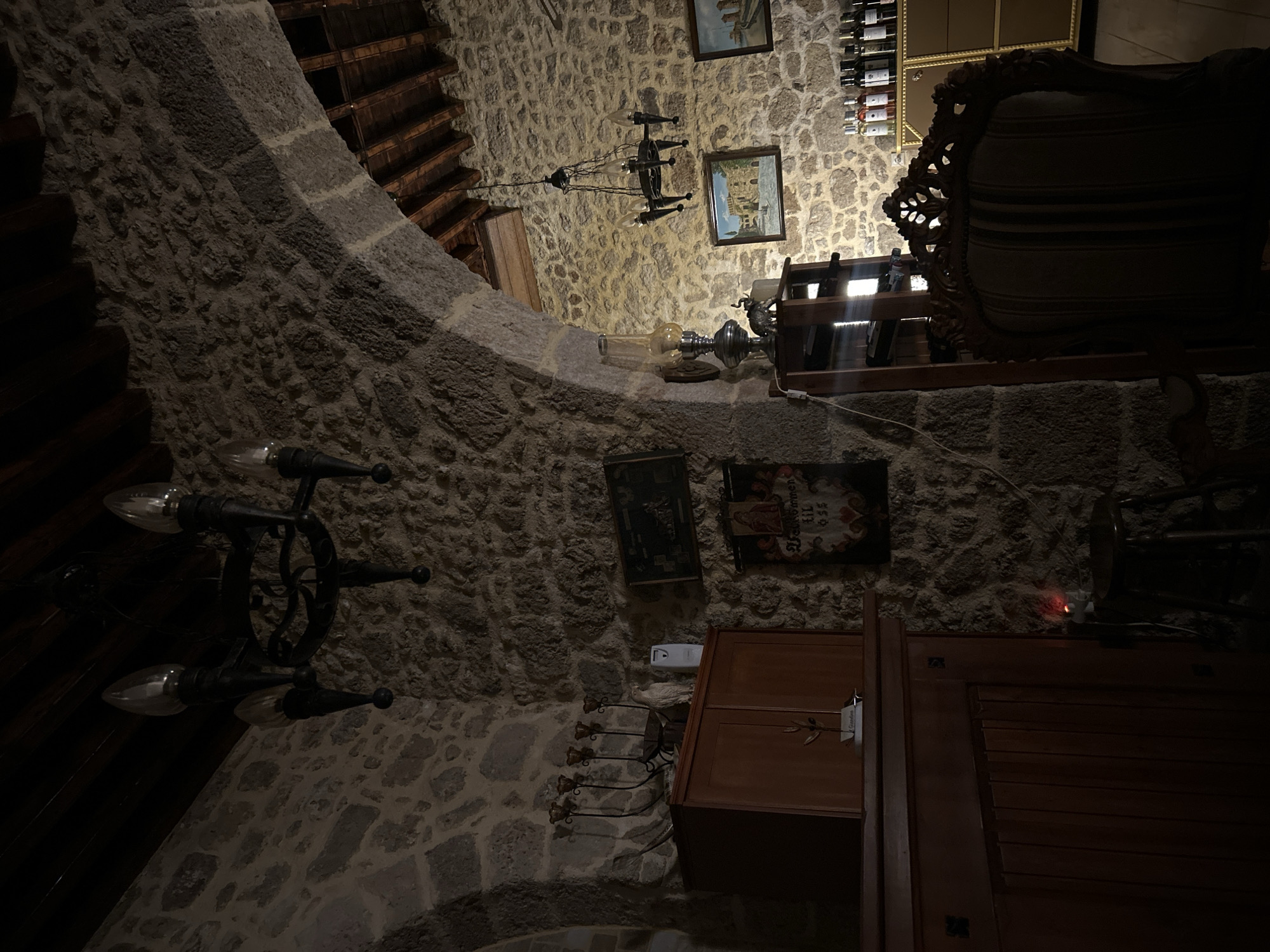 The image depicts the interior of a rustic stone building, possibly a restaurant or a historic home, located in the Municipal Unit of Rhodes, Greece. The scene is dimly lit, creating an atmospheric setting.
Dominating the upper left of the frame is a dark, wrought-iron chandelier with multiple arms, each holding a faux candle bulb. The stone walls are rough-hewn and irregular, characteristic of ancient architecture. An arched stone doorway or passage is visible, leading to another area with wooden ceiling beams.
On a wooden cabinet to the left, a candelabra with five faux candle holders is present, along with a white bird figurine and a small white appliance. Above the cabinet, a dark framed picture hangs on the stone wall. Below this, a banner with red and black designs and text, possibly in a foreign language, is displayed.
To the right of the arched passage, a vintage oil lamp with a metal base and glass shade sits on a wooden shelf unit, which also holds bottles of wine. A decorative elephant figurine is placed near the lamp. Further to the right, on the wall, are two framed landscape paintings. Below these paintings, a tall, ornate golden cabinet is visible, stocked with bottles of wine.
In the foreground, a dark, upholstered armchair with intricate carvings on its frame is positioned. The overall impression is one of historical charm and a cozy, dimly lit ambiance. No people are visible in the image.