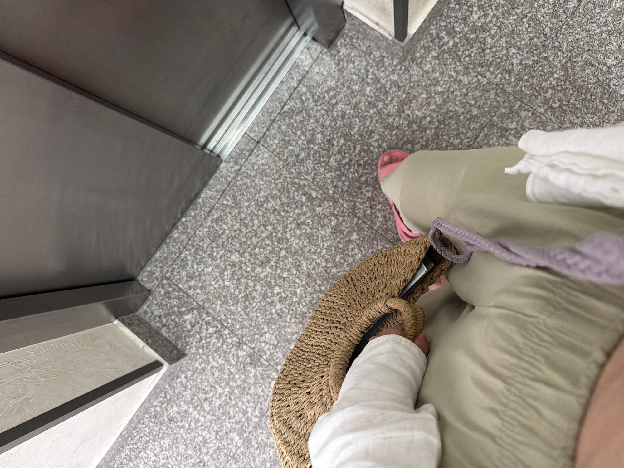 An overhead shot shows a person's legs and torso, seated near an elevator. They are wearing a light-colored top and light-colored pants. A straw bag rests on their lap. A pink sandal is visible on their right foot. The floor is a speckled gray tile. To the right, the stainless steel elevator doors are partially open. The wall to the left is textured and light-colored. The scene appears to be inside a building in the city of Budva, Montenegro.