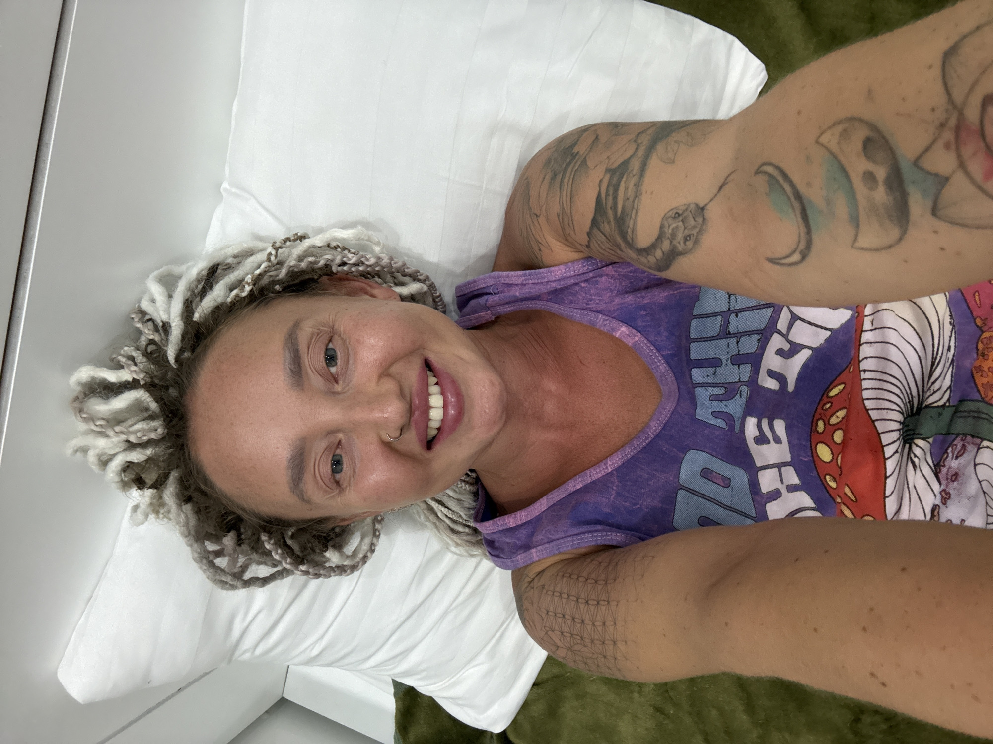 A woman with blonde dreadlocks and a purple tank top is lying on a bed, looking at the camera. She has a nose ring and her arms are covered in tattoos. The bed is covered in a white sheet and a green blanket. The scene appears to be indoors, possibly in a hotel room. The image is a selfie taken from a slightly high angle. The woman is smiling and appears relaxed.