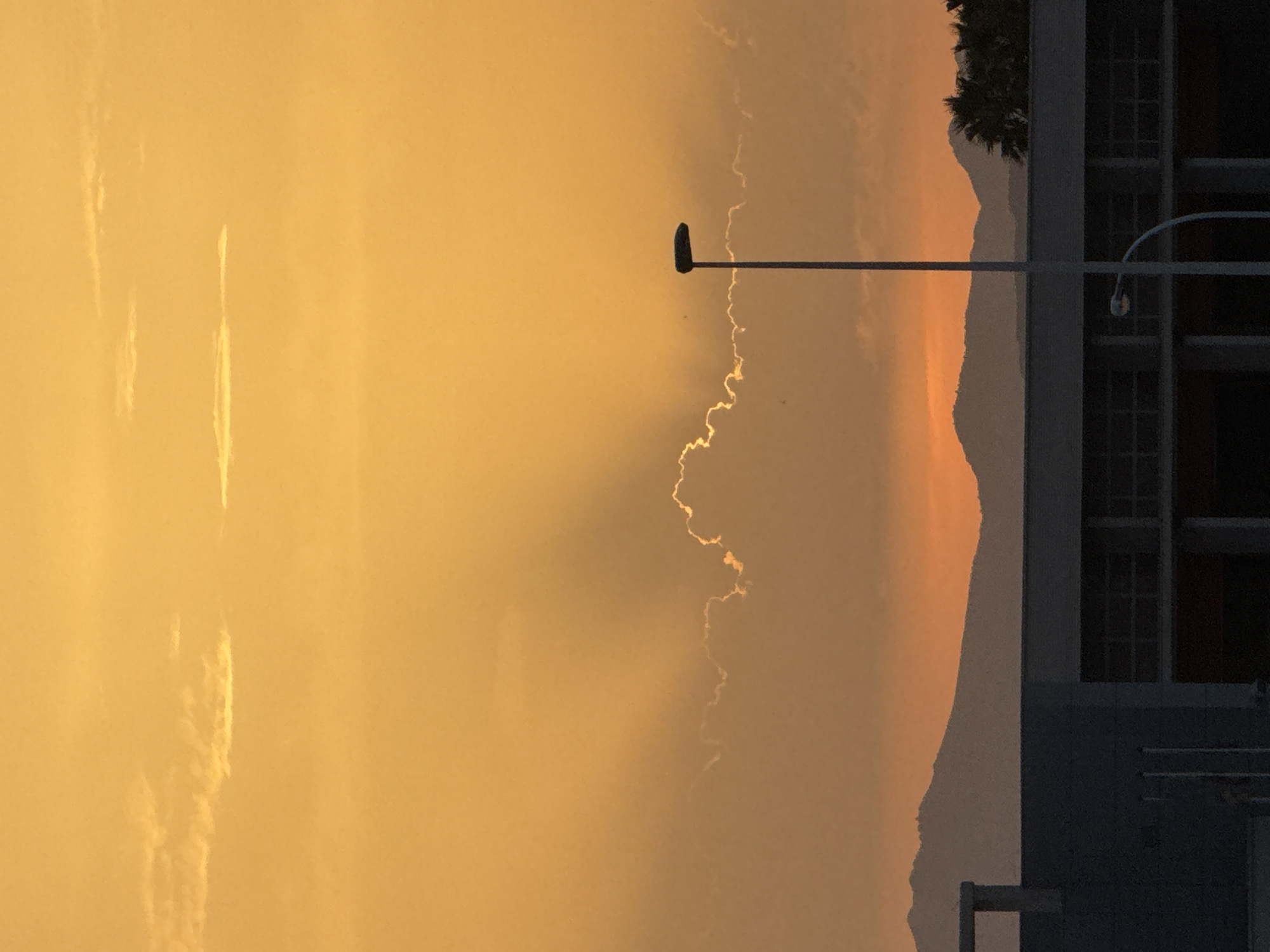 The image captures a twilight scene in Torrevieja, Spain, dominated by a vibrant orange sky. Silhouetted mountains form the distant horizon. In the foreground, a modern building with a dark facade and large windows is visible. A street lamp stands prominently in the middle ground, its modern design contrasting with the natural landscape. Wisps of clouds, illuminated by the setting sun, create dramatic light rays across the sky. The overall atmosphere is serene and captures the beauty of the coastal city at dusk.