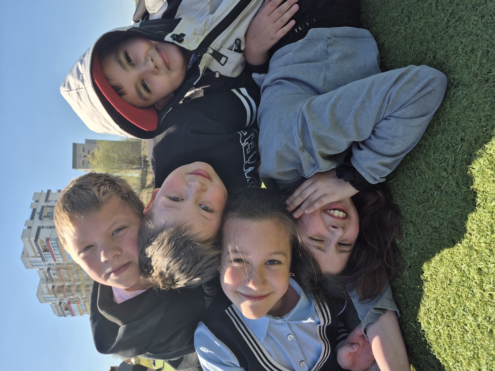 A group of five children are lying on artificial grass outdoors. The scene appears to be located in Chaikovsky, Russia, as indicated by the architectural style of the high-rise residential buildings in the background, visible in the upper-left corner.
From the bottom to the top of the image, a girl with long dark hair, dressed in a light blue collared shirt and a dark vest, is smiling directly at the camera. To her left, a boy with short brown hair and fair skin wears a dark hoodie. To his left, a boy with light brown hair looks at the camera with a neutral expression.
Above the three children, a boy with black hair and a dark hoodie smiles gently. Further up, another child wearing a white hooded jacket looks downward.
To the right of the first two children is a girl with long dark hair, wearing a grey long-sleeved shirt, covering the right side of her face with one of her hands and smiling brightly.
The children appear to be arranged in a circle with their heads together. The background consists of artificial grass, and beyond that, there are residential buildings and a clear blue sky. The lighting suggests it is daytime.