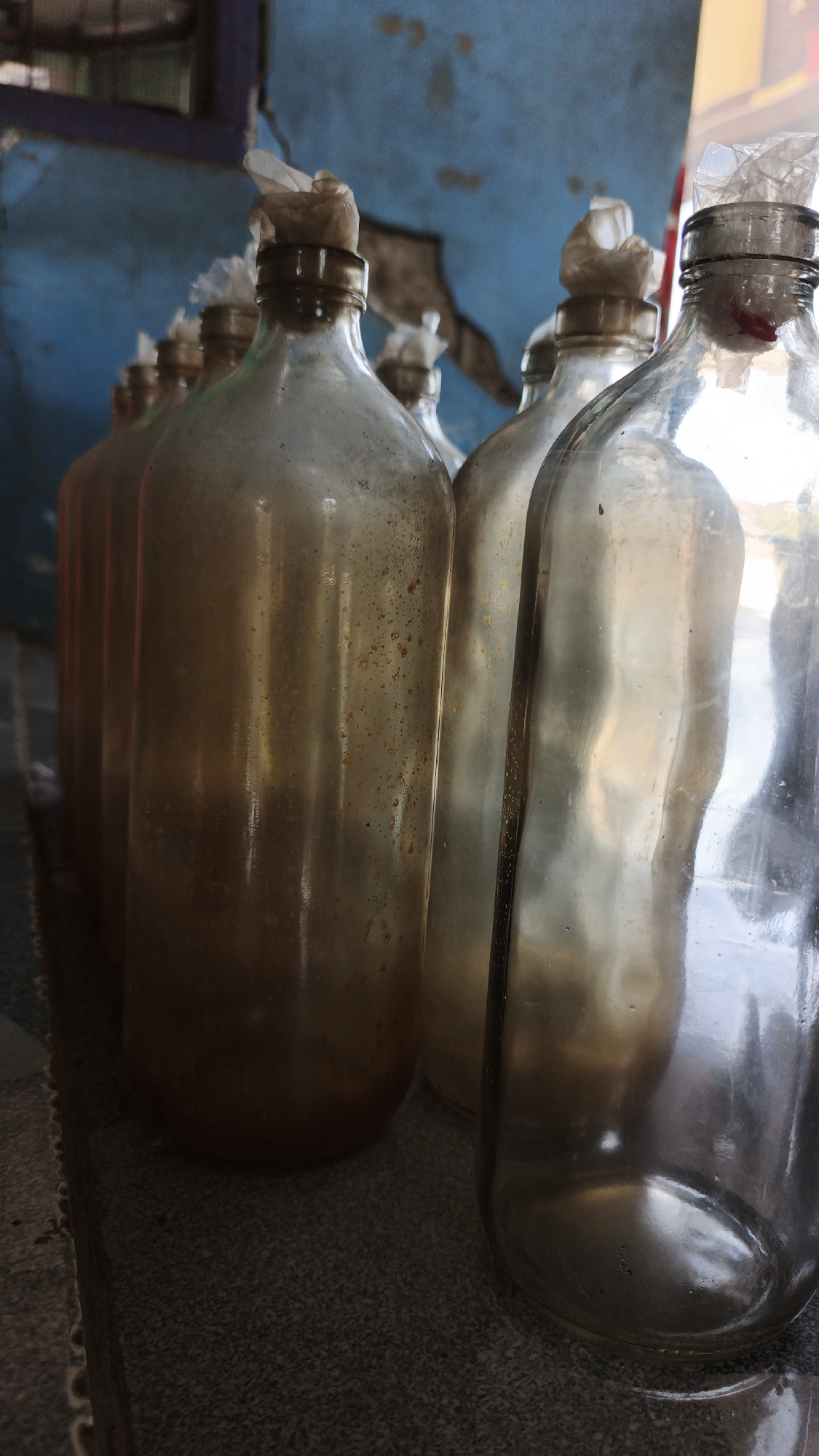 The image shows a row of glass bottles with plastic wrapped around their necks. The bottles are old and dirty, with some liquid at the bottom of the first few bottles in the row. The background is a blue wall with some peeling paint. The bottles are arranged on a surface that appears to be concrete. The scene is likely in a storage area or a place where these bottles are being reused or prepared for disposal. The specific location in Santing, Indonesia is not evident from the image itself, as there are no identifying landmarks or context clues. The image is a close-up shot, focusing on the details of the bottles and their condition.