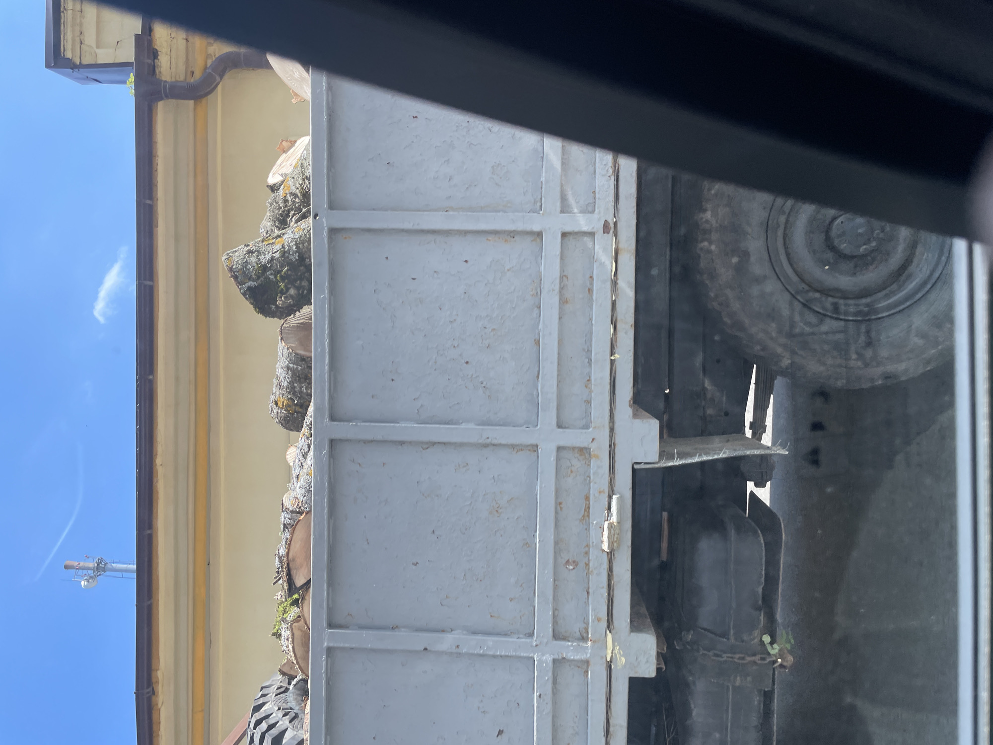 The image shows the side of a gray truck bed filled with logs and debris, viewed from a slightly low angle. The truck is parked in front of a building with a yellowish facade and a brown-edged roof. A dark drainpipe is visible on the right side of the building. In the background, a clear blue sky with a few wispy clouds is visible, along with a tall cell tower. The truck's tire and part of its undercarriage are also visible. The scene appears to be an outdoor setting, likely in a city given the building and cell tower. Without specific identifying landmarks, it is not possible to determine the exact location within Stavropol, Russia.