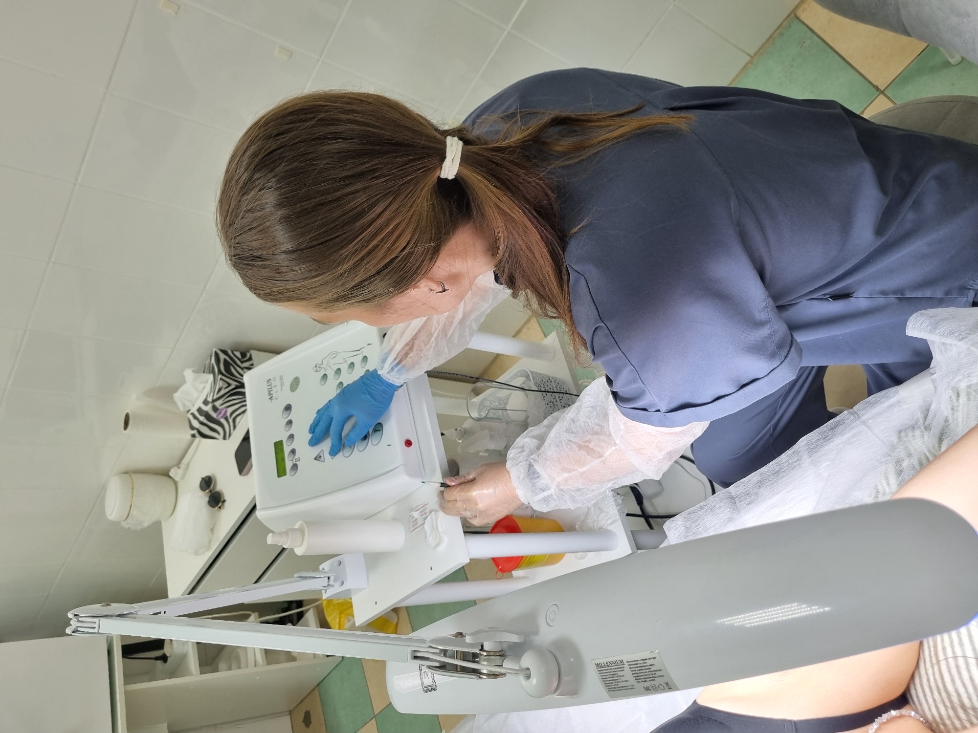A female cosmetologist, wearing a blue scrub top, blue gloves, and a white disposable gown, operates a medical device in a clinic. The device, labeled "APILUS CLEO," features a digital display and buttons, which she is interacting with. Her other hand, also gloved and in a disposable sleeve, holds a pen-like instrument over a white surface with cotton pads. The clinic environment includes a white equipment cart with various bottles, a yellow biohazard bin, and a bright adjustable lamp. In the background, there is a white cabinet, a roll of paper towels, a tissue box with a zebra print, and sunglasses. A patient, partially visible in the lower left, is lying on a striped surface, with a grey device extending over their body. The flooring is tiled in a green and white checkered pattern. The scene suggests a cosmetic or dermatological procedure is underway. The location is specified as a clinic in Chaikovsky, Russia.