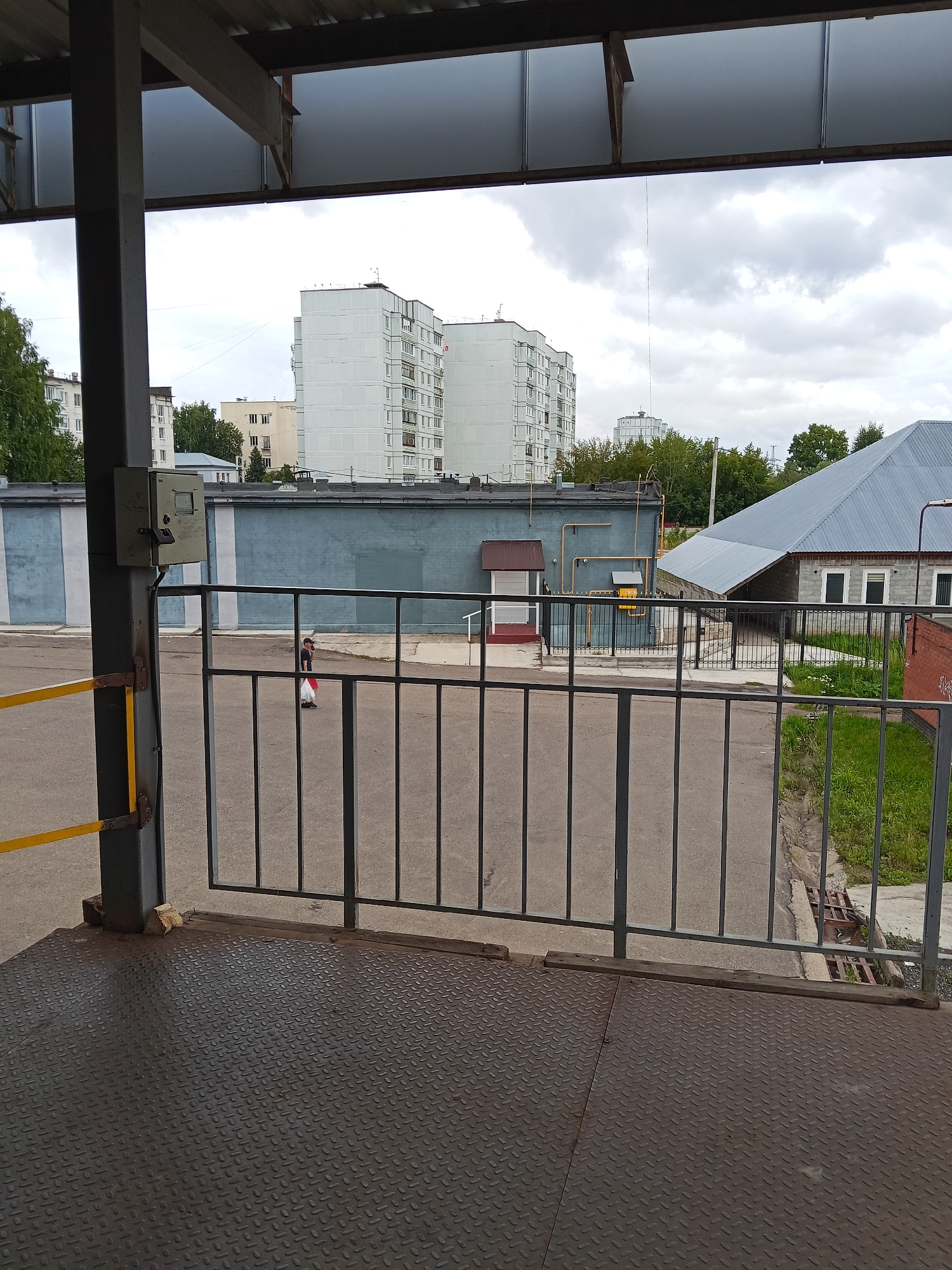 The image depicts an outdoor scene likely in Chaikovsky, Russia, viewed from under a covered area with a metal roof and support beams. In the foreground is a dark, textured metal platform. Beyond this, a grey metal gate with vertical bars separates the viewer from a paved area.
In the middle ground, a person wearing a dark top and white pants, carrying a white bag, is walking away from the camera towards the right. Behind the gate and the person, a long, low blue building with grey stripes is visible. To the right of this building is a grey-roofed structure with brick walls and white-framed windows, and further to the right, some green grass and a brick wall.
In the background, several tall, white apartment buildings with many windows dominate the skyline under a cloudy sky. Trees are also visible between the buildings. A yellow and black railing is attached to a dark grey support column on the left side of the image, near a metal box mounted on the column. The overall environment appears to be an urban or industrial area with residential buildings in the distance.