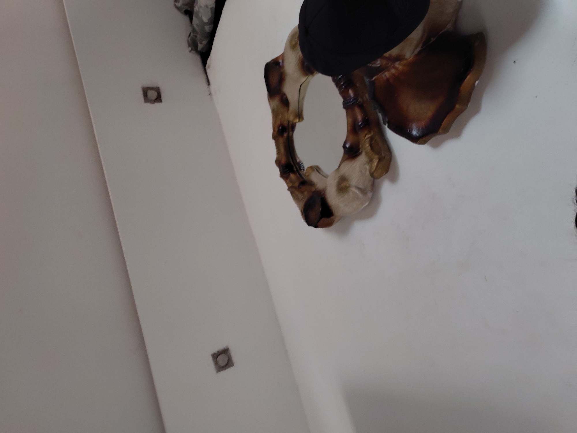 The image displays an indoor scene, likely a white wall in a room, though the original image is tilted. A distinctive, ornate mirror is centrally featured on the white wall. The mirror's frame has an organic, irregular shape, resembling twisted wood or roots, and exhibits a rich color palette of dark browns, golden-browns, and light cream or beige tones, suggesting it might be made of carved wood or a similar natural material. Part of a black object, possibly a hat or a piece of dark clothing, is visible above the top portion of the mirror frame.
Additionally, two square, recessed fixtures are visible on the white wall. These appear to be either small light fixtures or electrical outlets/boxes, positioned at different points across the wall. No specific activity or event is taking place; the image is a static shot of an interior space. The time of day and weather are not discernible, and there is no visible text within the image. The scene is characterized by its clean white background contrasting with the warm, earthy tones of the mirror frame. The location context is Santa Ana, Venezuela, though no specific visual cues within the image indicate this.
