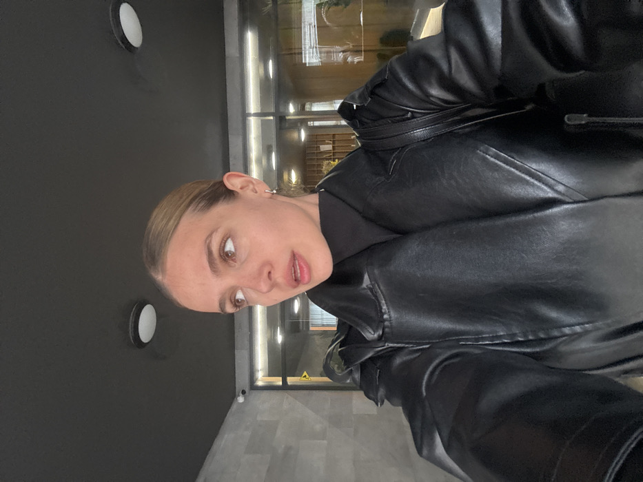 An overhead-angle selfie captures an adult female from the chest up, positioned in an indoor hallway or lobby. She has fair skin, light brown hair pulled back from her face, brown eyes, and her mouth is slightly agape. She wears a black turtleneck garment under a black faux leather trench coat or jacket, and a silver hoop earring is visible on her left ear. A black shoulder bag strap rests on her left shoulder. Her gaze is directed upwards and to her left, off-camera.
The background features a dark gray ceiling with two circular, recessed white light fixtures. A small, dark security camera is mounted on the ceiling in the upper left quadrant. The wall to the left appears to be light gray and textured. The wall to her right is primarily a glass partition, revealing an interior space with warm lighting, blurred architectural elements, and indistinct green foliage. A yellow triangular sign with a black symbol is faintly visible through the glass on the left side of the glass partition. The location is an indoor modern building entrance or common area, city of Unknown, Unknown.