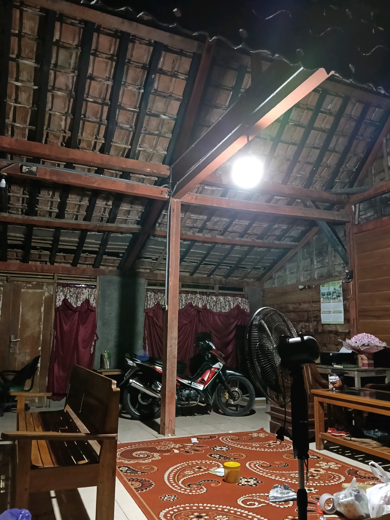 The image shows the interior of a room with a tiled roof and exposed wooden beams. A motorcycle is parked inside the room, with a large standing fan positioned in front of it. A wooden bench is visible in the foreground, and a patterned rug covers part of the floor. To the right, a wooden table holds a bouquet of pink flowers. A calendar with the text "ANDA JALAN" is mounted on the wall. The scene appears to be a residence in Galuk, Indonesia. There are no people visible in the image.