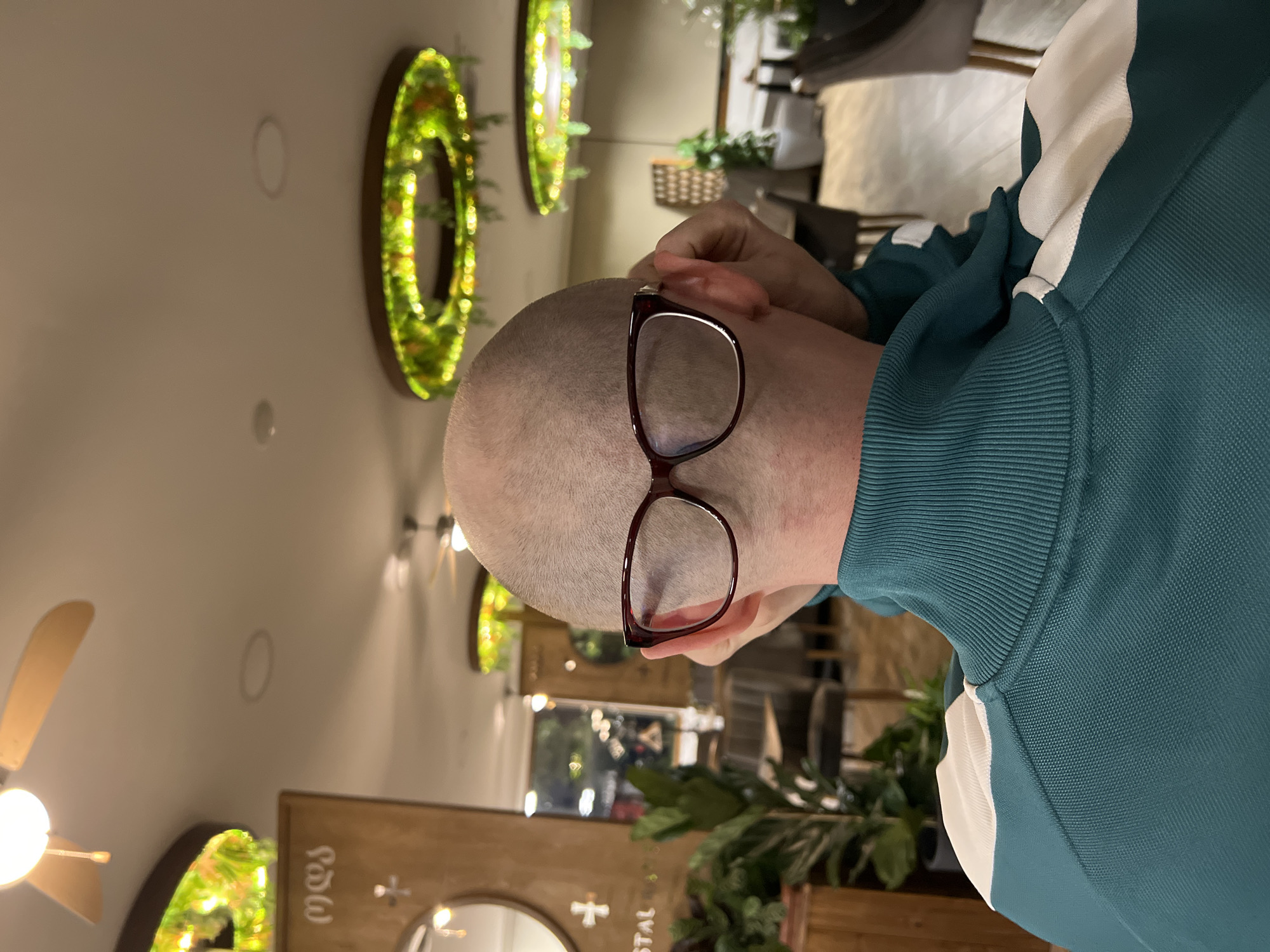 A person with a shaved head and wearing glasses is seen from behind, partially obscured by the right shoulder and arm wearing a dark teal and white tracksuit. The person appears to be in an indoor setting, potentially a restaurant, based on the decor and furniture visible in the background.
Behind the person are several decorative elements:
* Circular green wreaths with embedded lights hanging on a light-colored wall.
* A partially visible wooden frame with what appears to be Georgian script and a cross design.
* Various indoor plants.
* Other indistinct furniture and decor elements suggestive of a commercial or public setting.
There is no direct evidence within the image to confirm that the location is Boreti, Montenegro. It is impossible to know the location from just the image.