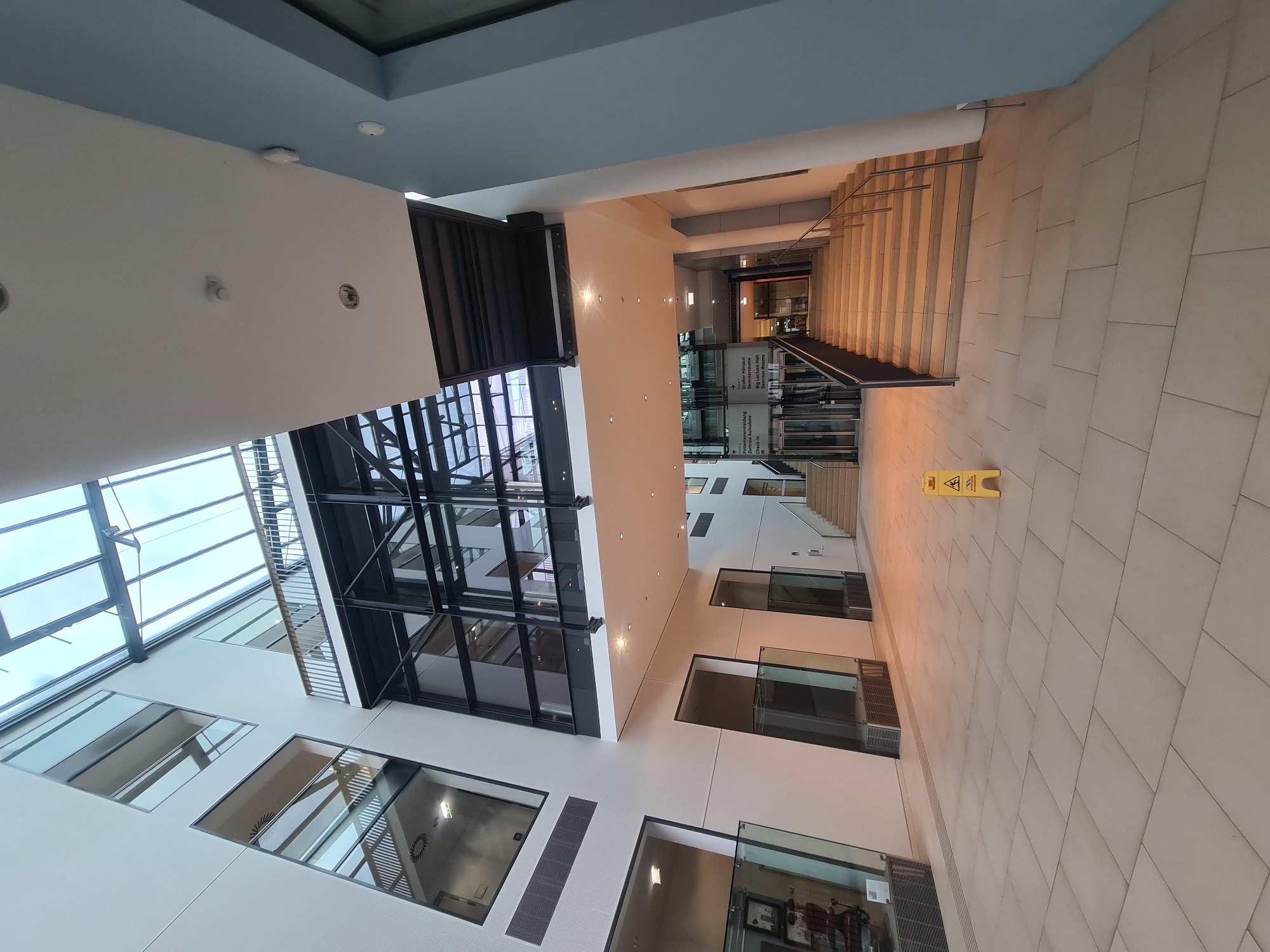 The image depicts the interior of a modern building, likely a public space or office, with a large atrium. The architecture features a multi-story design with prominent glass elements, including a glass ceiling and internal windows. A central staircase leads upwards, flanked by railings. To the left, there are glass-enclosed rooms or offices, and a separate set of glass doors. A yellow caution sign, indicating a wet floor, is visible on the light-colored tiled floor in the foreground. Signs with German text are present near the elevators and staircase, one of which reads "Patiëntenanmeldung Zentrale Aufnahme" (Patient Registration Central Admission) and another indicating "Großer Hörsaal Seminarräume" (Large Lecture Hall Seminar Rooms). The lighting appears to be a combination of natural light from the ceiling and artificial interior lights. The overall environment is clean and contemporary, suggesting a well-maintained institutional or commercial setting. Based on the German signage, the location is likely within a German-speaking country. While no specific landmarks are visible to pinpoint Vienna, Austria, the presence of German signage makes it a plausible location within Austria.