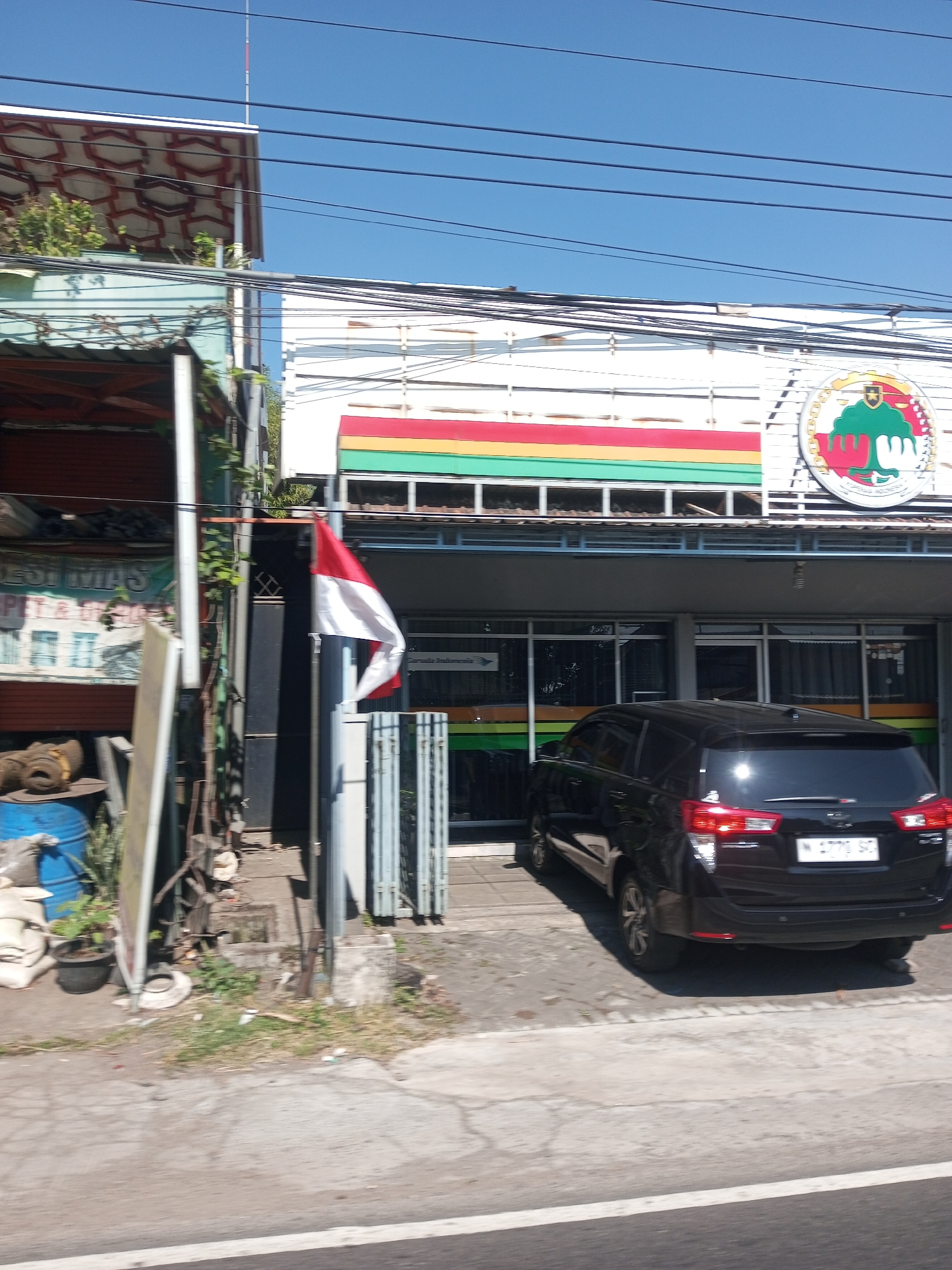 The image captures a street scene in Sengon Agung, Indonesia, on a clear, sunny day. In the foreground, a black SUV is parked in front of a building. The building appears to be a shop or business, with a prominent sign featuring horizontal stripes of red, yellow, and green, and a circular emblem with a green tree. An Indonesian flag, red and white, is planted on a pole to the left of the building's entrance. Adjacent to the building on the left is another structure with an open-fronted area displaying various items, including large sacks and a blue barrel. The street is paved, with a white line marking the edge of the road. Overhead, power lines crisscross the sky. The environment suggests a typical urban or semi-urban setting in Indonesia.