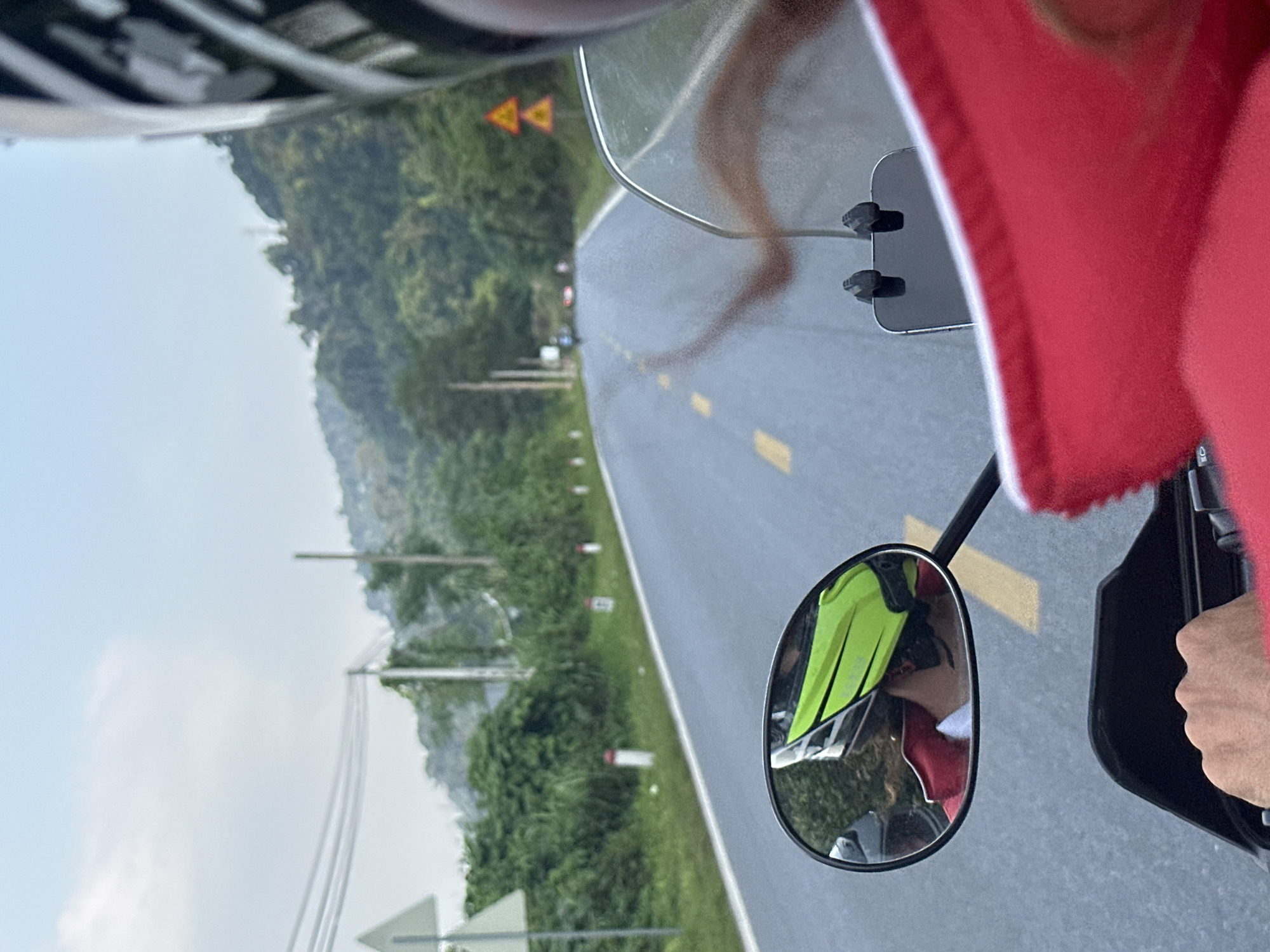 The image captures a first-person perspective from a motorcycle rider or passenger traversing a paved, winding road in a mountainous and verdant landscape, likely in Vị Xuyên Commune, Vietnam.
In the foreground, elements of the motorcycle and rider are visible. The top-left corner shows a part of a helmet with a black and white pattern. On the right, a red garment, possibly a jacket or sun protection, is prominent, and a hand firmly grips the motorcycle's handle. A clear windshield reflects a blurry, elongated shape that appears to be legs. A rearview mirror on the right offers a reflection of the person's torso, clad in a bright neon yellow vest or jacket over a red layer, confirming the presence of at least one person on the bike.
The main subject is the road ahead, stretching into the distance. It is a well-maintained asphalt road with distinct yellow dashed lines marking the center. The road curves gently through a landscape dominated by lush green mountains and hills covered in dense vegetation under a bright, clear sky. Utility poles with multiple power lines run parallel to the road on the left side, and white and red roadside markers are intermittently visible. In the mid-distance on the left, two yellow triangular warning signs with red borders are posted, featuring black symbols that indicate upcoming road hazards, likely curves or winding road sections.
The scene suggests an outdoor journey or road trip, likely during daylight hours given the bright illumination and clear visibility. The weather appears fair and sunny. The winding road and mountainous terrain align with the known characteristics of Vị Xuyên Commune, offering scenic views. No specific emotions are discernible, but the setting conveys a sense of travel and exploration.