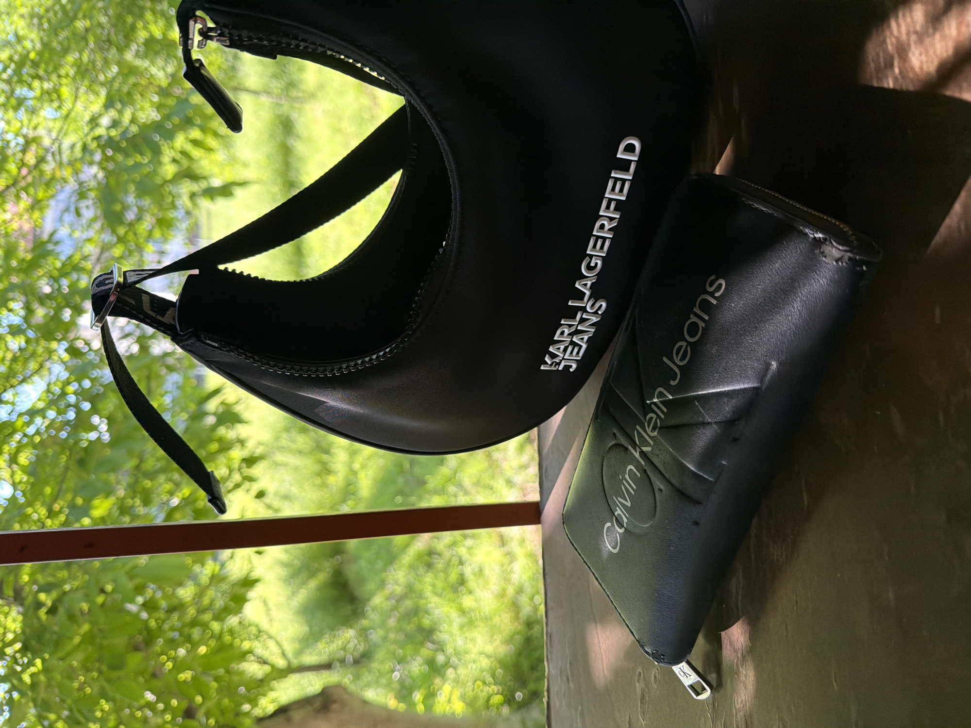 The image shows a black Karl Lagerfeld Jeans handbag and a black Calvin Klein Jeans wallet on a weathered surface, likely outdoors, based on the presence of green foliage and dappled sunlight. The handbag is crescent-shaped with a visible shoulder strap. The wallet appears to be a zip-around style. The objects are situated near a metal or wooden structure, with greenery blurred in the background. This photo was taken in Troyan, Bulgaria.
