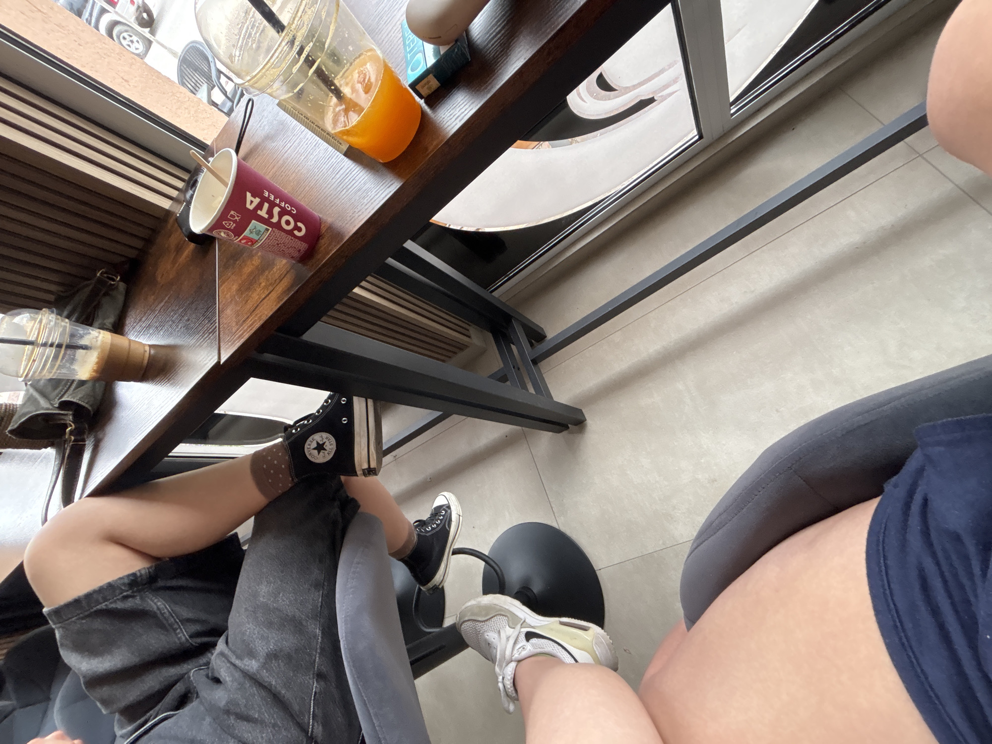 The image depicts a high-angle shot of a table and two people sitting at a cafe. On the table, there are two plastic cups - one contains an orange liquid and the other a dark red one with the word "COSTA" written on it. One person is wearing grey denim shorts and black Converse sneakers and brown polka dot socks. The other person is wearing navy shorts and light-colored sneakers with white laces. The setting appears to be indoors, possibly at a café with natural light coming in from the right side of the frame. The location in Garmen, Bulgaria is not discernible based on the visual elements of the provided image.