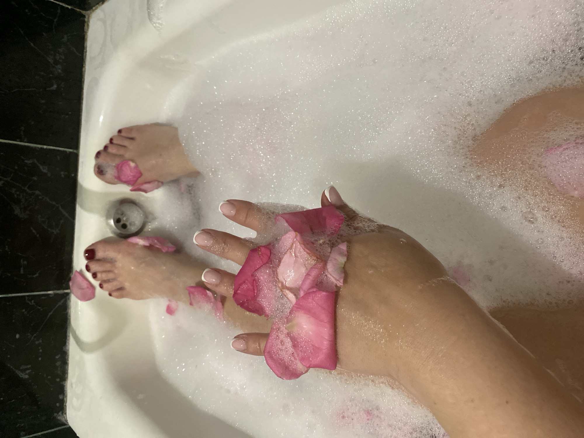 The image depicts a partial view of a person in a bathtub filled with bubbly water and adorned with rose petals. The person's feet and hands are visible, with rose petals scattered on them. The feet have dark red nail polish. The hand has a French manicure. The bathtub is white, and there are black tiles visible near the edge of the tub. The location is specified as Sochi, Russia, but there are no visual clues within the image to confirm this location; it appears to be a bathroom.
