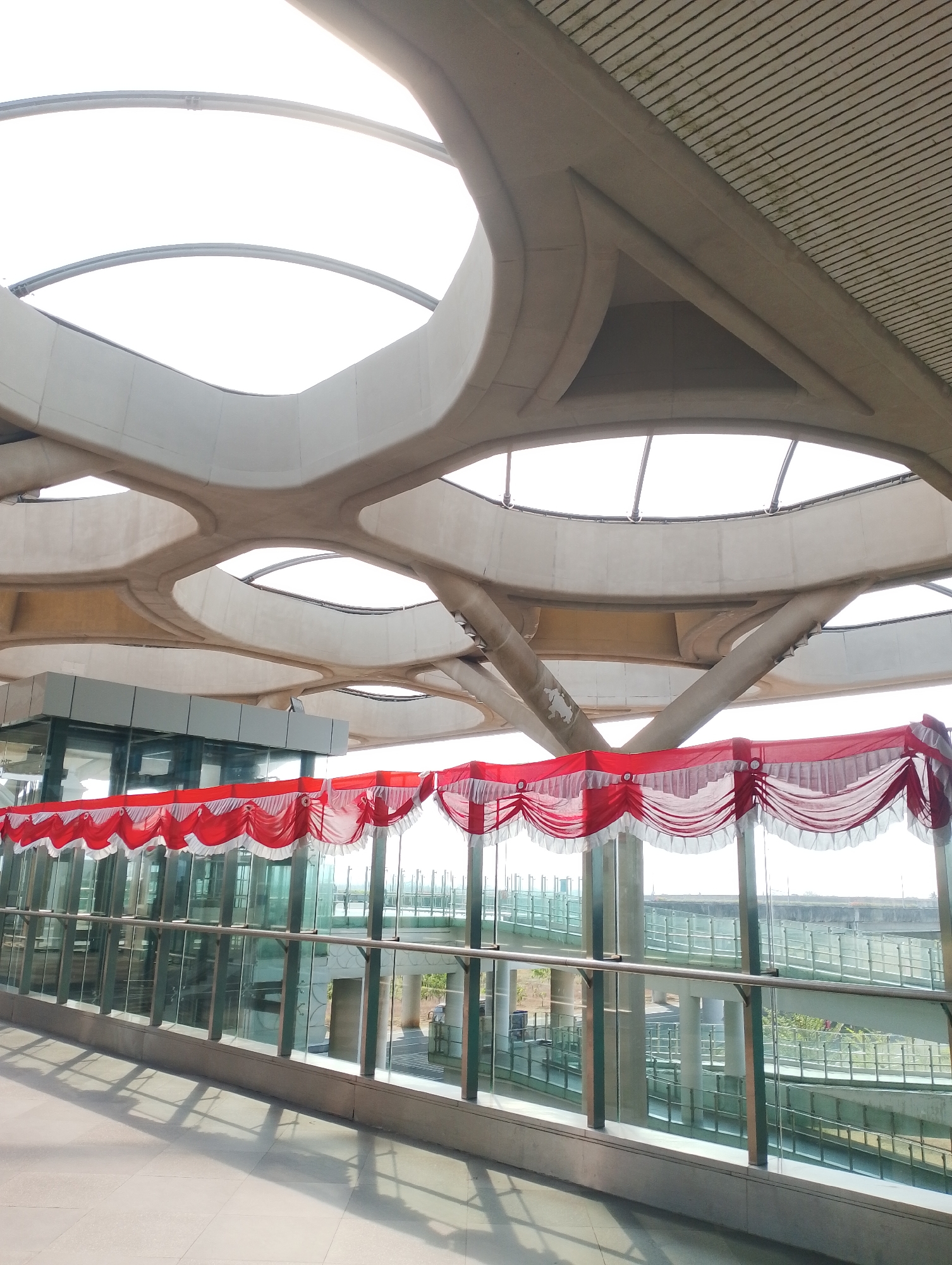 The image shows a modern architectural interior with a distinctive curved ceiling structure and large windows. Red and white decorative fabric is draped along the top of the windows. Through the windows, elevated walkways and structures are visible, suggesting an airport or transportation hub. The flooring is tiled. The overall impression is one of a clean, contemporary public space. The image is likely taken in the public area of the Yogyakarta International Airport in Palihan, Indonesia.