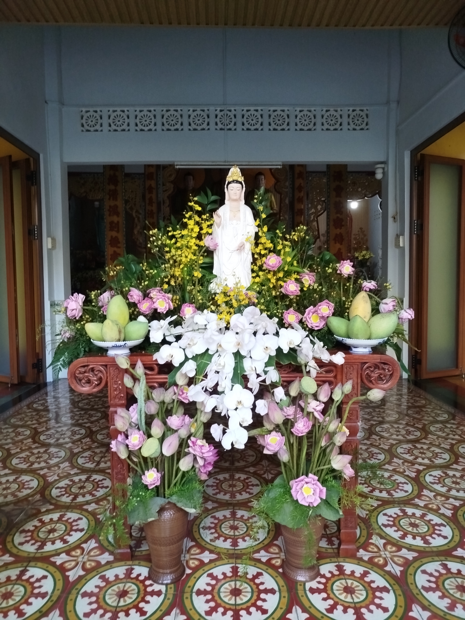 A statue of a Buddha-like figure stands on a wooden table, surrounded by an abundance of flowers in various colors including white, pink, and yellow. The table also holds a bowl filled with green fruit, possibly mangoes. The statue is positioned against a backdrop of ornate wooden panels, potentially depicting religious scenes or motifs. The scene appears to be set within a building, possibly a temple or shrine, as suggested by the decorative flooring and the religious icon. Two doorways are on both sides of the table. The pattern on the tile is circular. It is in Ho Chi Minh City, Vietnam, based on the cultural setting and the statues of Buddha like figures.
