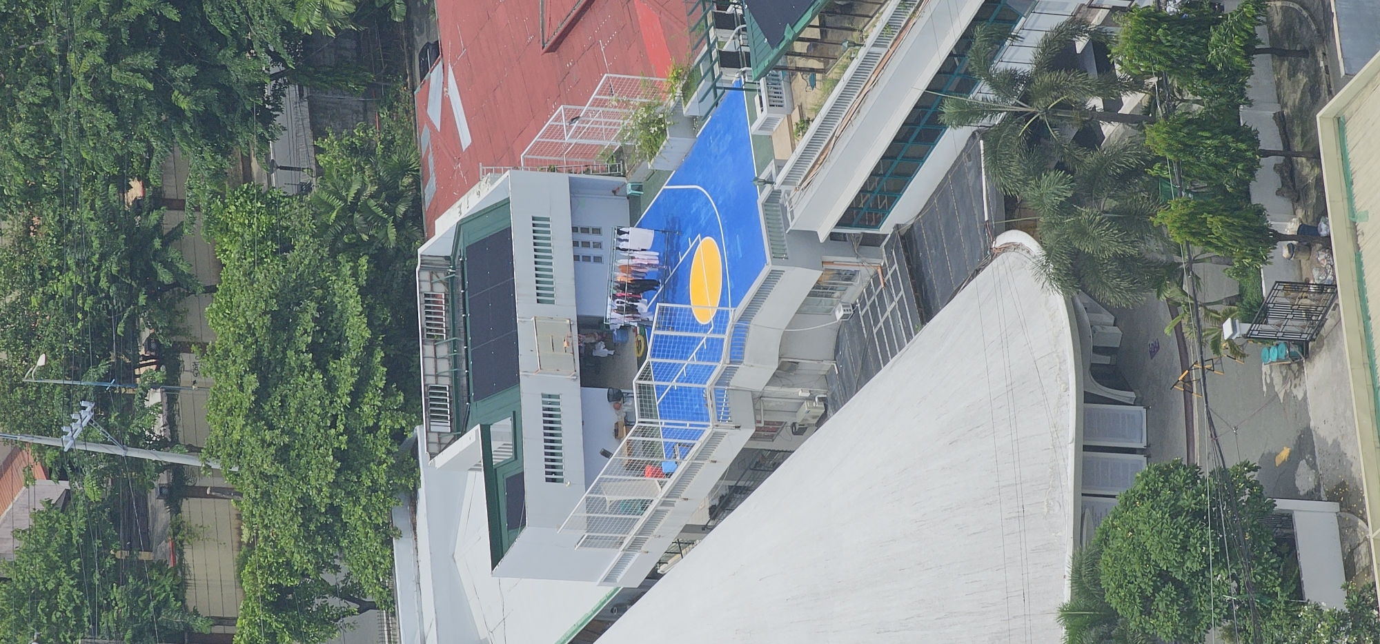 The image captures an aerial view of a building complex in Makati City, Philippines, featuring a rooftop basketball court. The court is painted bright blue with a yellow center circle. Several individuals are present on the court, and clothing is hung on a clothesline on the side. Adjacent to the court is a multi-story building with a white facade, featuring numerous windows. The roof of this building is partially covered with solar panels. Beyond this structure, another building with a red roof is visible, along with lush green trees surrounding the complex. The scene suggests an active community space within an urban setting.