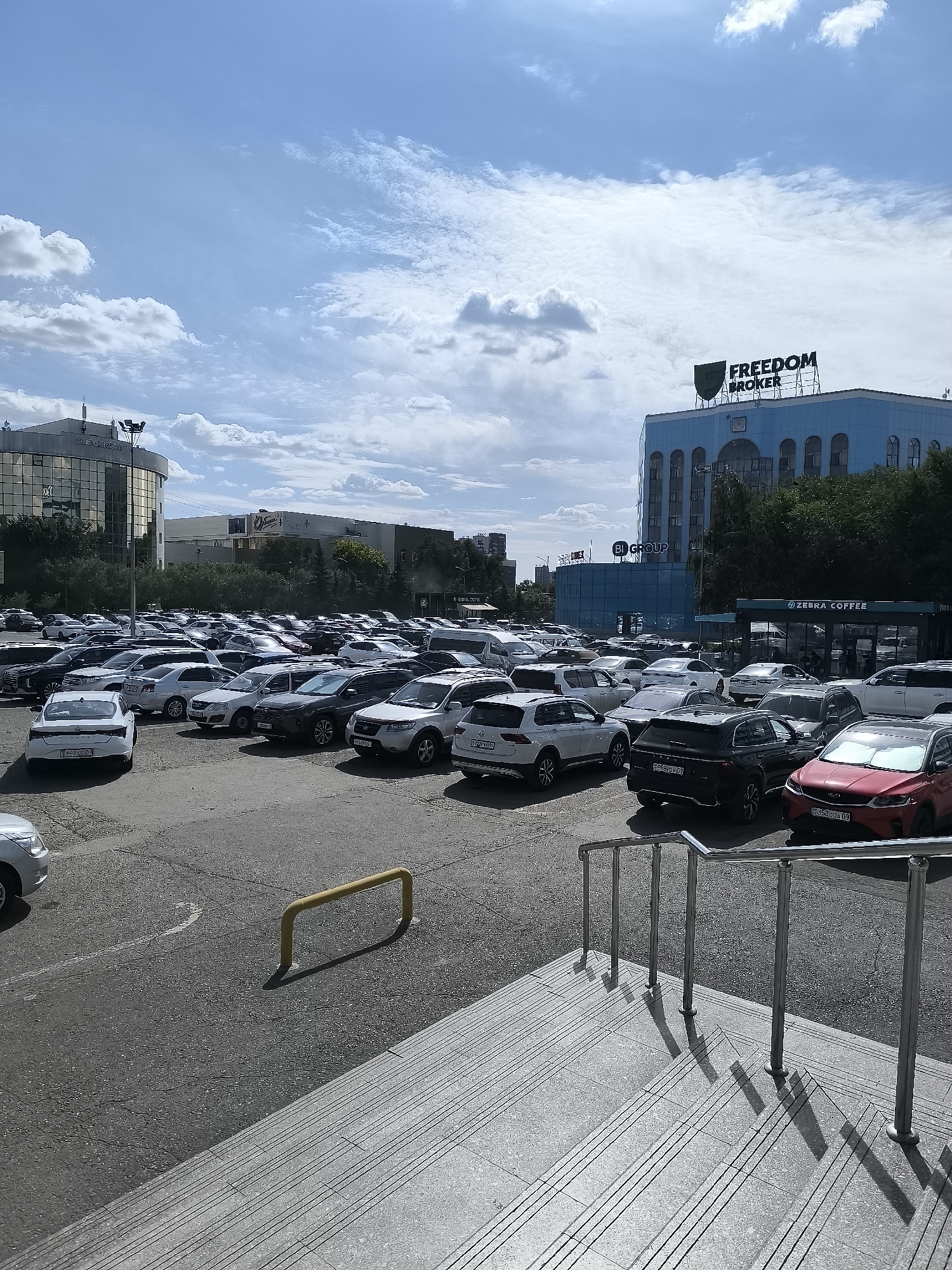 A wide-angle, eye-level, outdoor, daytime, full shot captures a vast, partially filled asphalt parking lot in front of a modern commercial building complex. The sky above is a mix of blue and white, with scattered clouds.
In the foreground, a gray concrete staircase with metal railings and shadows stretches from the bottom right of the frame towards the parking lot. A yellow metal barrier is visible on the left side of the parking lot.
The parking lot itself is occupied by numerous cars of various colors and models, parked in rows. The vehicles include sedans, SUVs, and hatchbacks. Some cars are white, black, gray, and red.
Behind the parking lot, a large, light-blue building with arched windows dominates the mid-ground. A prominent sign on top of this building reads "FREEDOM BROKER." To the left of the blue building, a taller, glass-fronted building with a curved facade is visible, with a sign that appears to read "AGROCREDIT." Further to the left, a narrower building can be seen.
On the right side of the blue building, a smaller structure with a dark awning and the sign "ZEBRA COFFEE" is present, with tables and chairs visible through the windows. Trees and foliage are present behind these buildings, indicating an urban setting.
The scene suggests a commercial area, possibly a shopping center or office complex, in the city of Qarağandı, Kazakhstan. No people are clearly visible in the parking lot or on the stairs, but the presence of parked cars and commercial establishments implies activity.