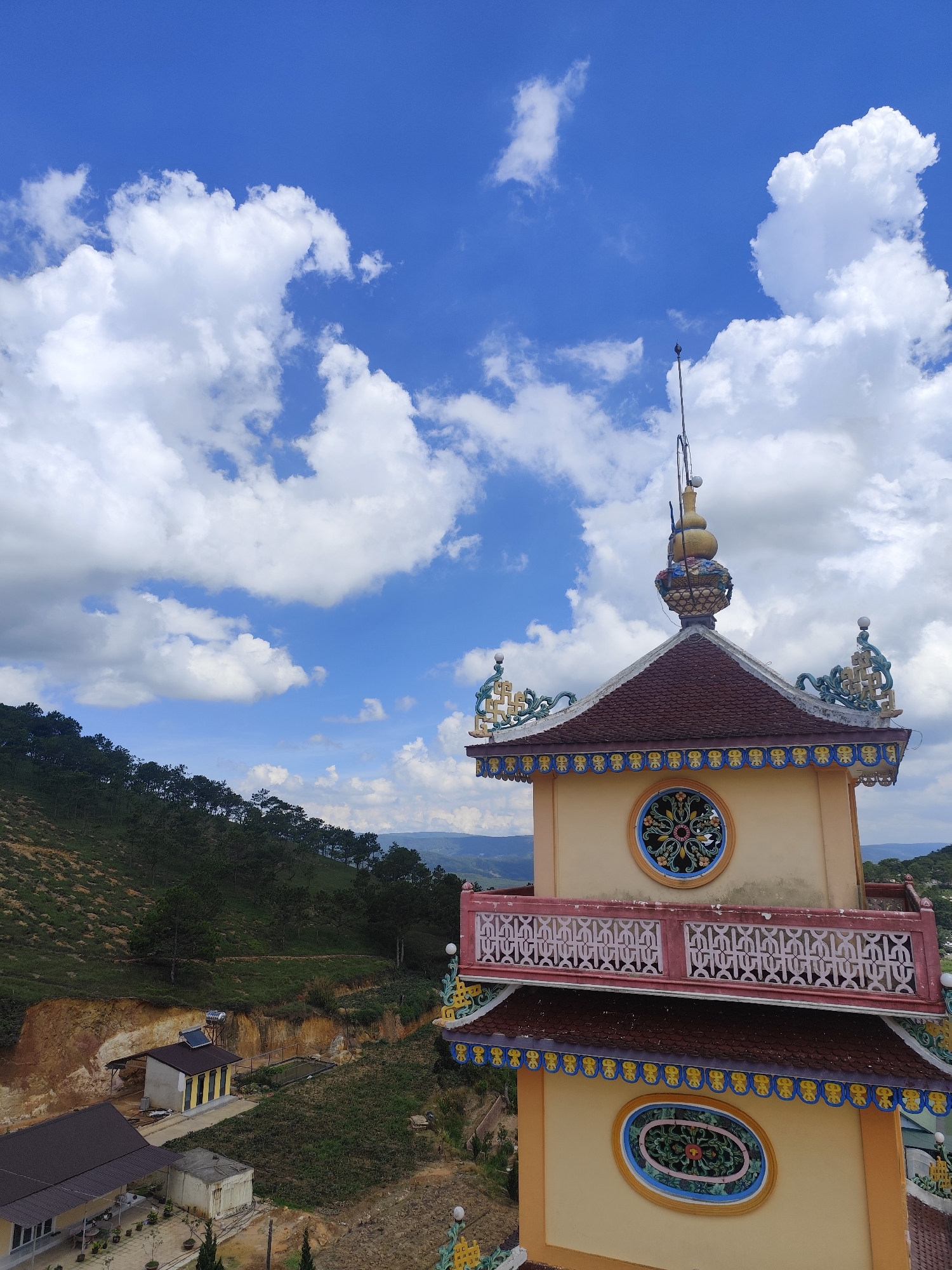 The image depicts a multi-story pagoda-like structure with ornate details, set against a backdrop of a lush green hillside dotted with trees. The pagoda features a red tiled roof with decorative yellow and blue trim. Circular stained-glass windows are visible on its facade. The sky above is a vibrant blue, adorned with fluffy white clouds. In the distance, rolling hills and mountains are discernible. In the foreground, to the left, part of a larger building with a dark roof is visible, along with a smaller structure featuring a solar panel. The scene is likely located in Lộc Quý, Vietnam, given the architectural style and the natural landscape. There are no people or animals present in the image, and no actions or interactions are occurring.