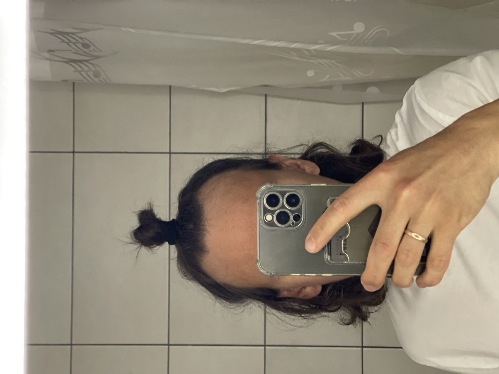 The image displays a close-up selfie of a person, likely taken in a bathroom in front of a mirror or reflective surface. The individual's head is visible, featuring long, dark brown hair pulled back into a small bun secured with a black elastic tie, and a noticeably receding hairline. The person is holding a smartphone with a clear protective case in their right hand, and a simple, golden-colored ring is worn on the fourth finger of that hand.
The background consists of light-colored square tiles with dark grout lines, characteristic of a bathroom. A shower curtain with a decorative, light grey pattern, possibly resembling stylized musical notes or waves, is reflected above the person's head. The lighting appears to be artificial, typical of an indoor setting.
Visible text on the phone case includes the word "GRIP" on an accessory, along with faint inscriptions such as "EMBOUSURE" and "DESIGNED FOR BLUE." The person's face is partially obscured by the phone, preventing a clear view of their expression. The time of day and external weather conditions are not discernible from the image.