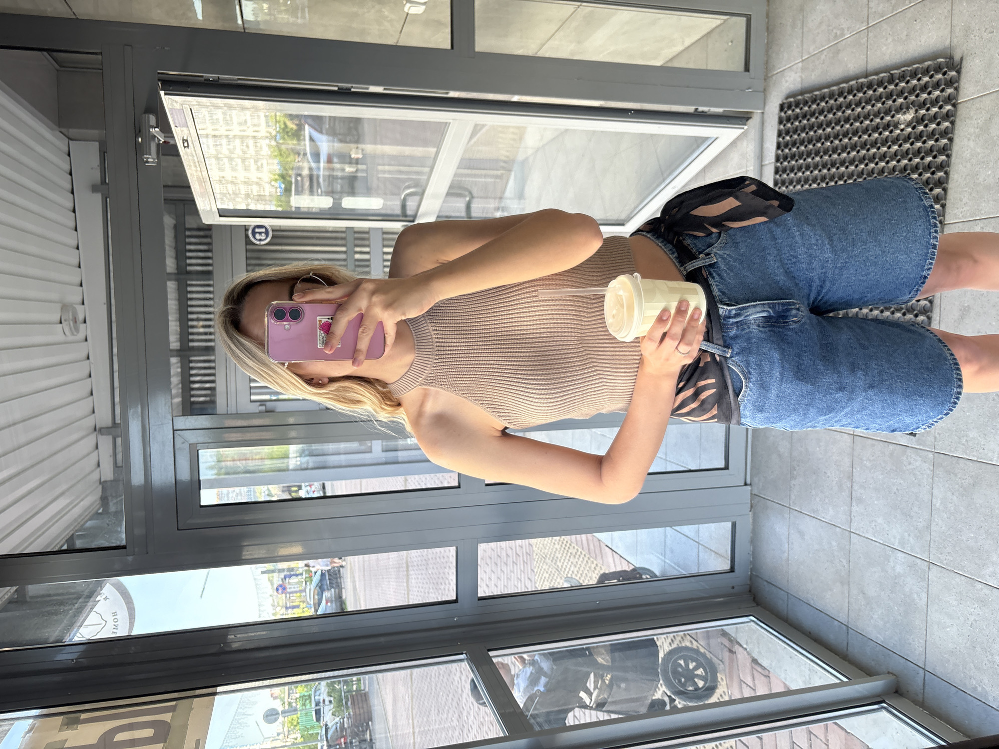 A young woman is depicted taking a mirror selfie indoors, likely within a building's entrance or corridor with large glass doors and windows. She is standing upright, holding a light pink iPhone with a sticker on the back in her left hand to take the photo, and a light yellow cup with a white lid and straw in her right hand. She wears a beige, ribbed knit halter top, blue denim shorts, and a black scarf with brown stripes tied around her waist as a belt. She has blonde hair, hoop earrings, and a ring on her right hand's ring finger.
The indoor setting features grey tiled flooring and a dark grey rubber mat near what appears to be an entrance. The glass surfaces extensively reflect the outdoor environment. Reflections reveal an urban street scene with buildings, vehicles (including a car and a baby stroller), and other details like a street sign with the number "13" in a circular graphic. A large, circular sign with partially visible text that appears to be "HOME" (or "ДОМ" in Cyrillic, considering the Krasnoyarsk, Russia context) is also reflected. The scene suggests daytime with bright, natural light.