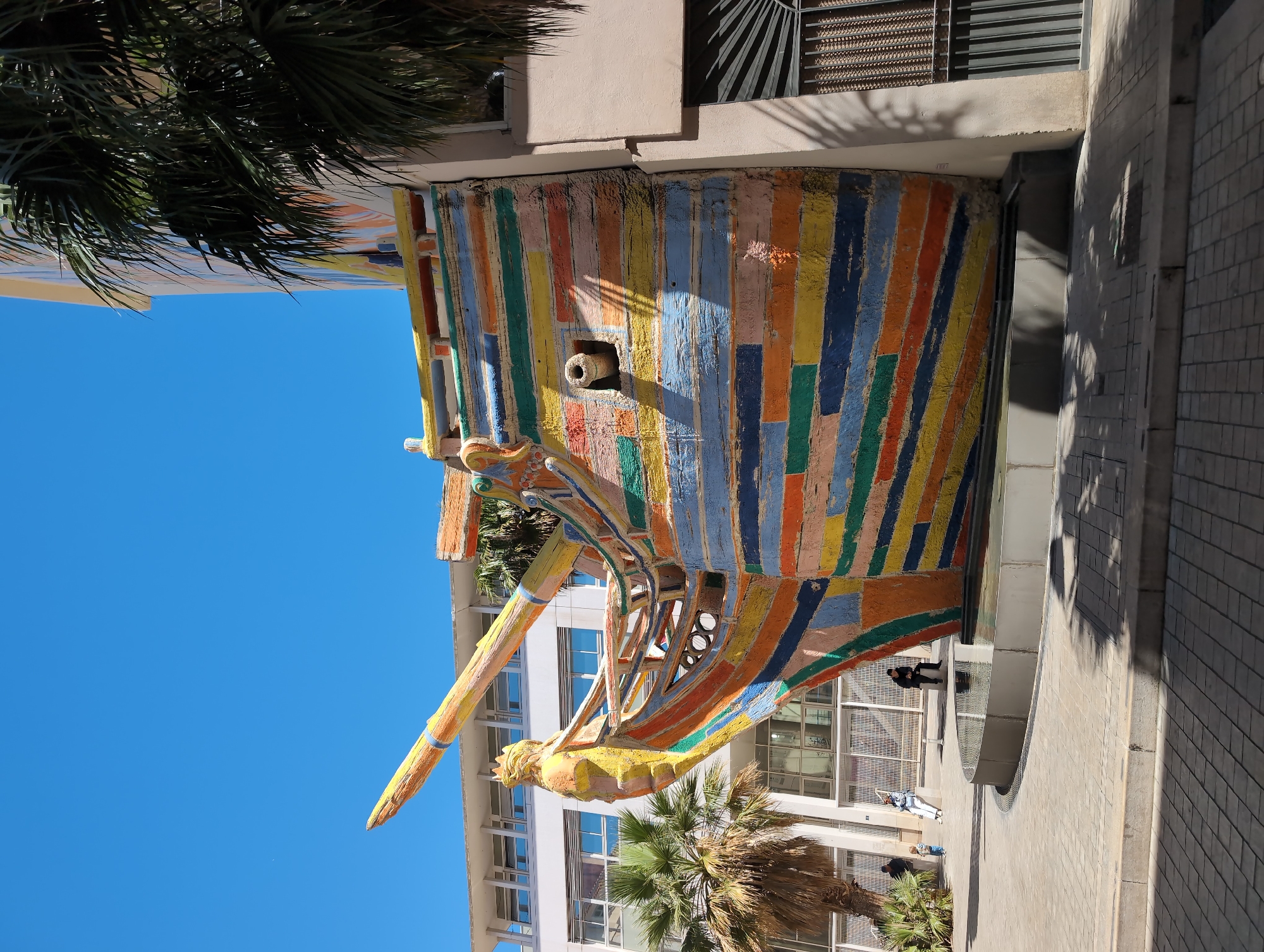 The image shows the "Fontaine du Dauphin" sculpture, a colorful, stylized prow of a ship, in Toulon, France. The sculpture is made of vertical planks of wood painted in bright colors like yellow, blue, orange, and green. A decorative figurehead adorns the front. Behind the sculpture is a modern building with white walls and multiple windows. In the background, a clear blue sky is visible. A palm tree stands next to the sculpture and the building. Pedestrians can be seen walking near the sculpture. The surrounding area appears to be paved with light gray bricks or stone.